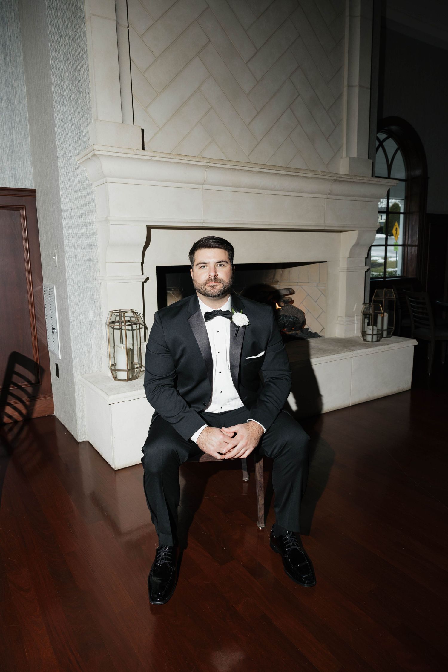 A groom in a black tuxedo sits casually against a white wall in a dramatic wide-angle shot.