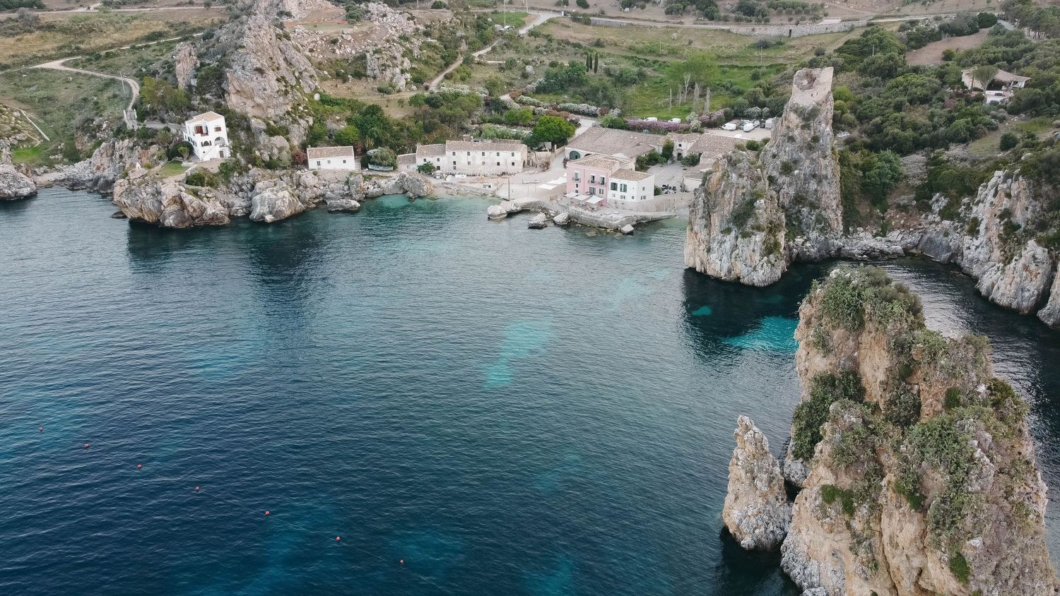 Wide aerial shot of a coastal settlement built into rocky cliffs overlooking turquoise Mediterranean waters.