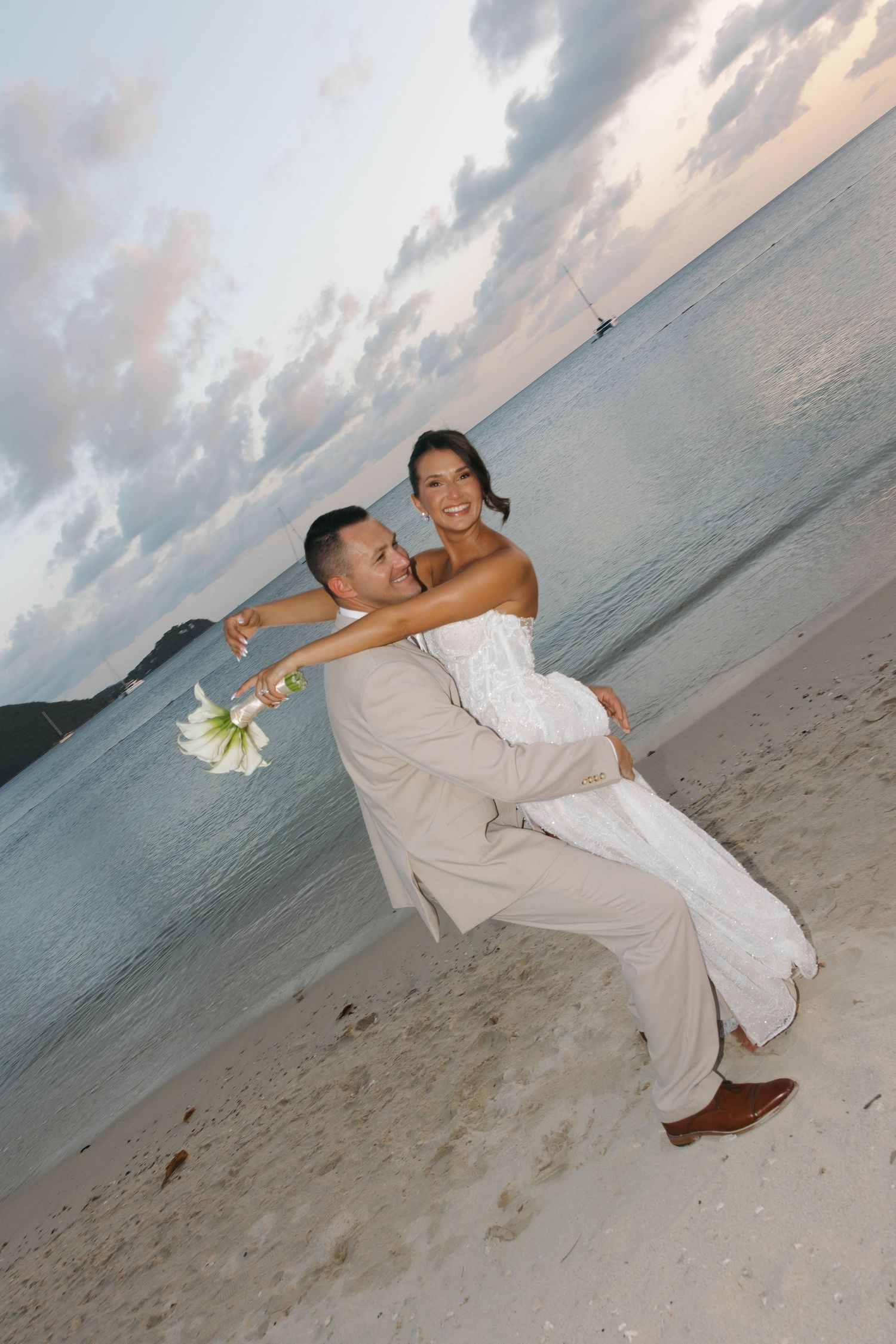 Newlywed couple celebrating on a tropical beach at sunset, groom lifting bride in white dress holding white flowers.
