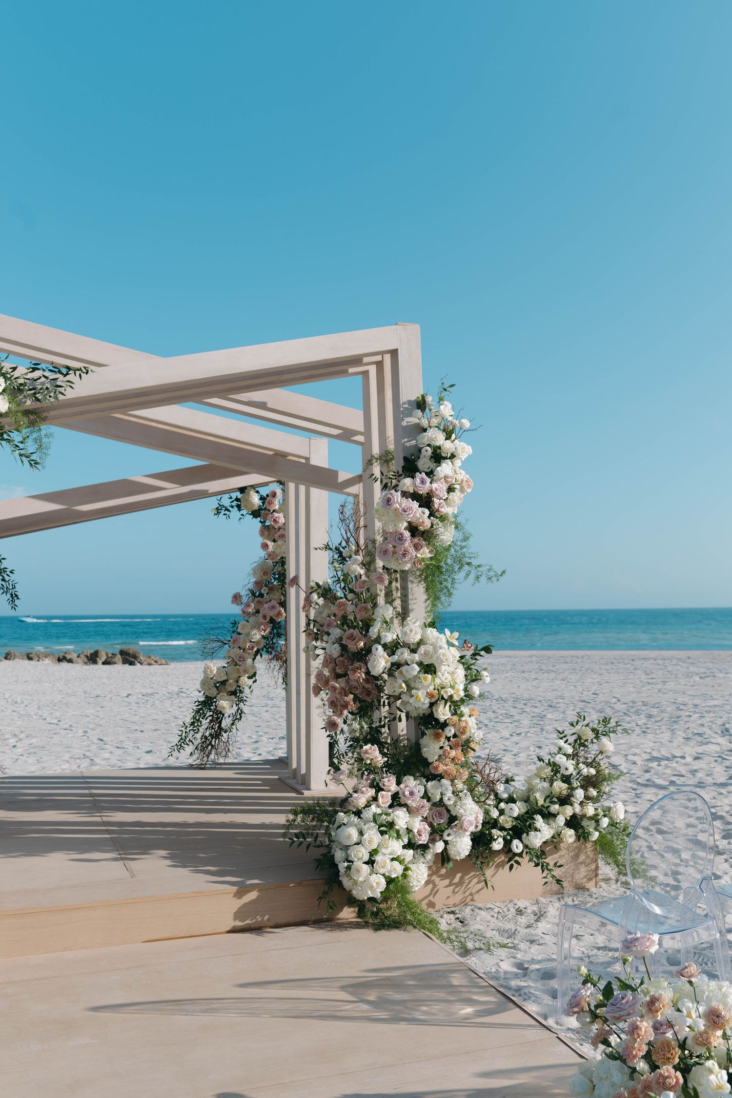 Modern geometric wedding arch with white and blush floral arrangements on sandy beach.