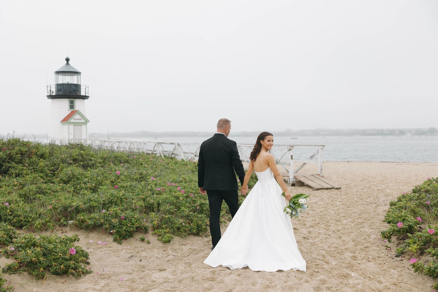 Wedding couple walks along coastal path with lighthouse in distance.