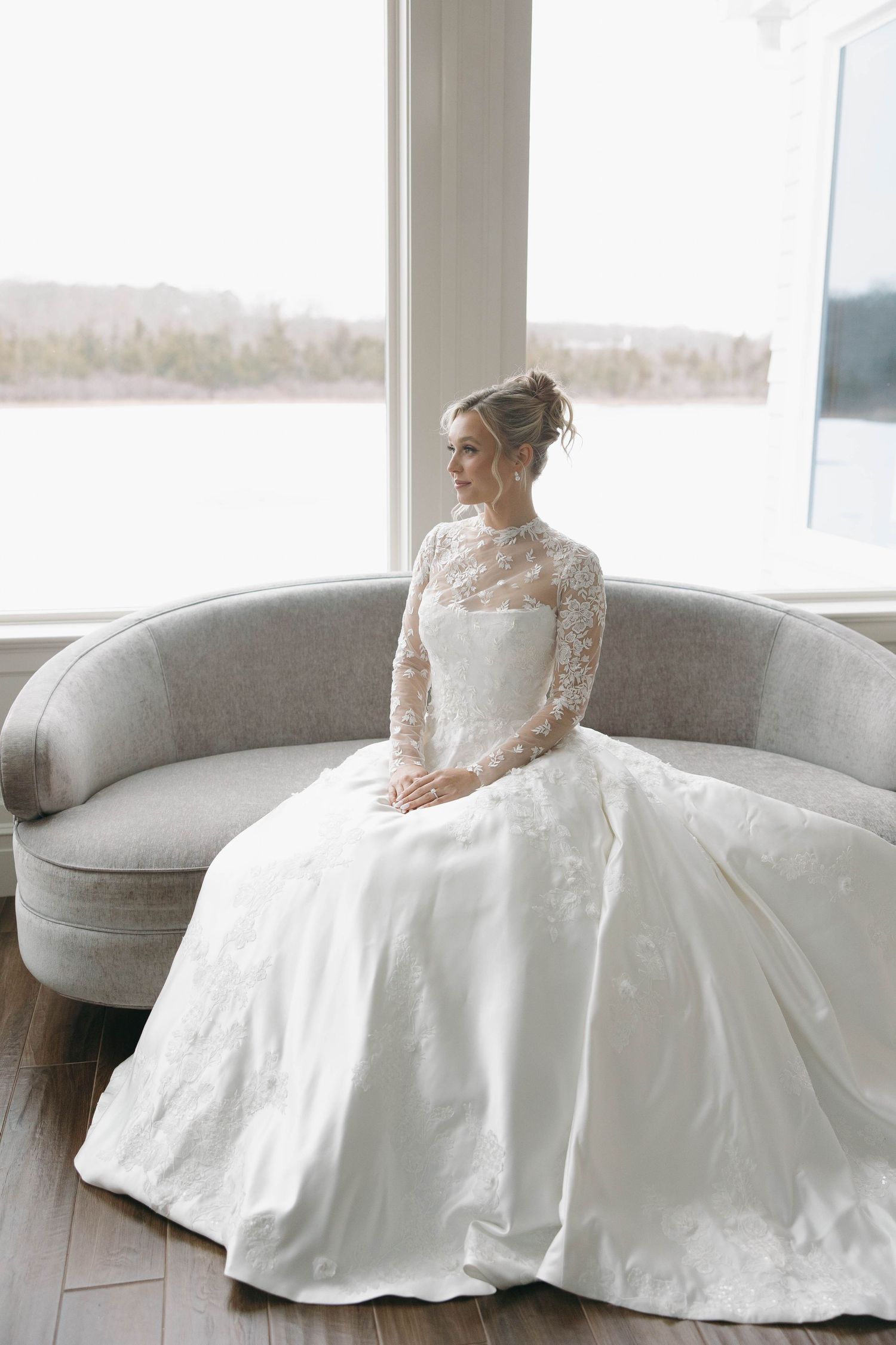 Wedding dress with full skirt displayed on grey curved sofa near bright windows.