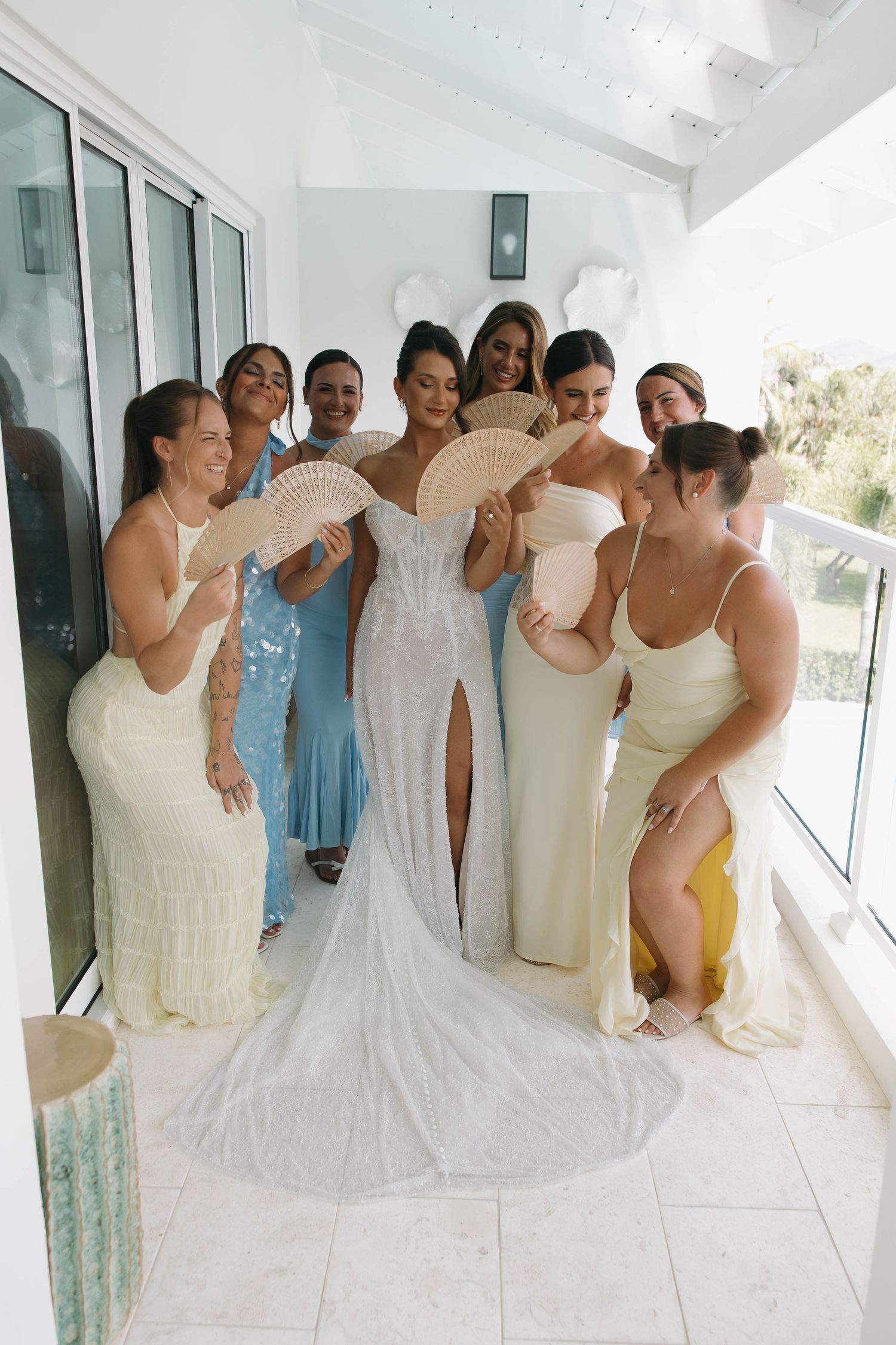 Bride in fitted lace wedding gown poses with bridesmaids holding fans on a bright white balcony.