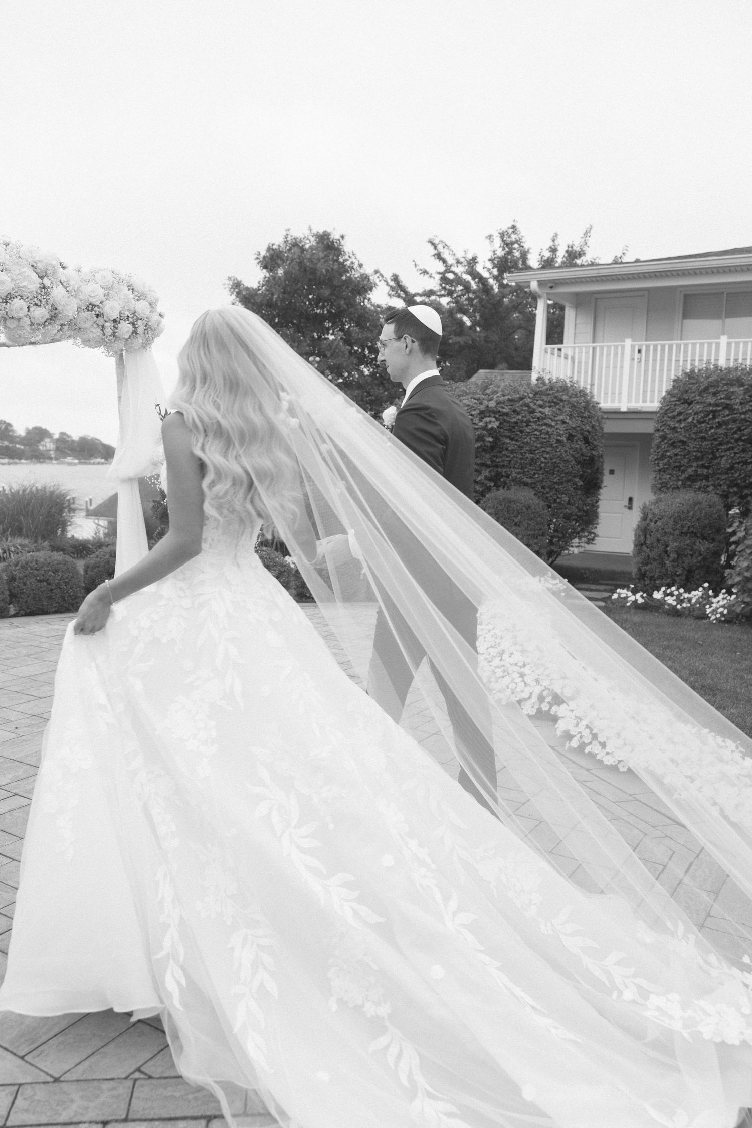 Black and white photo of a bride in a flowing lace gown and long veil standing with a groom outdoors.
