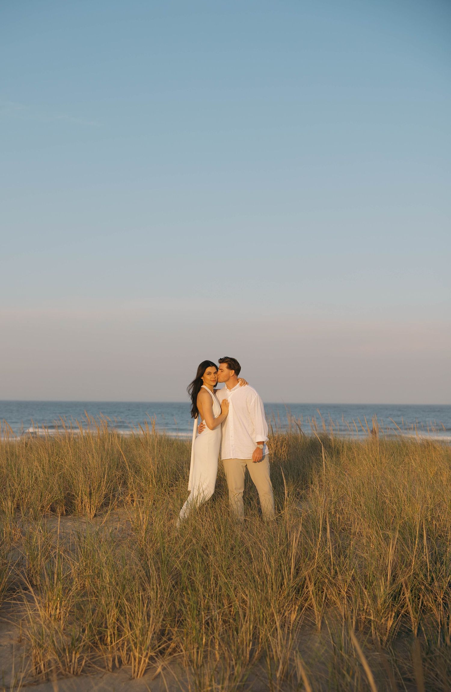 Couple in white attire sharing romantic moments among beach grass at sunset.