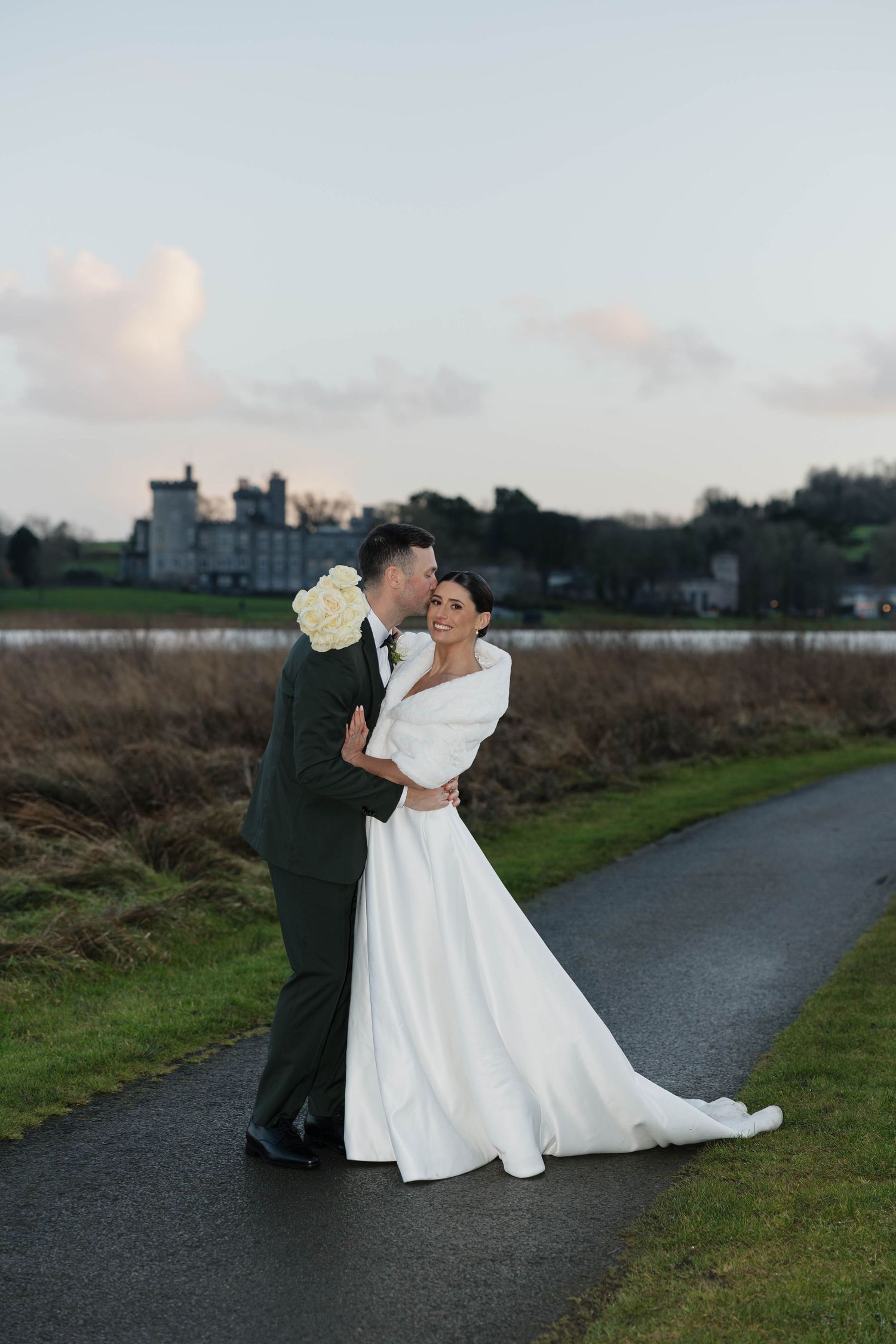 Bride in elegant white gown poses with groom on pathway near castle grounds.