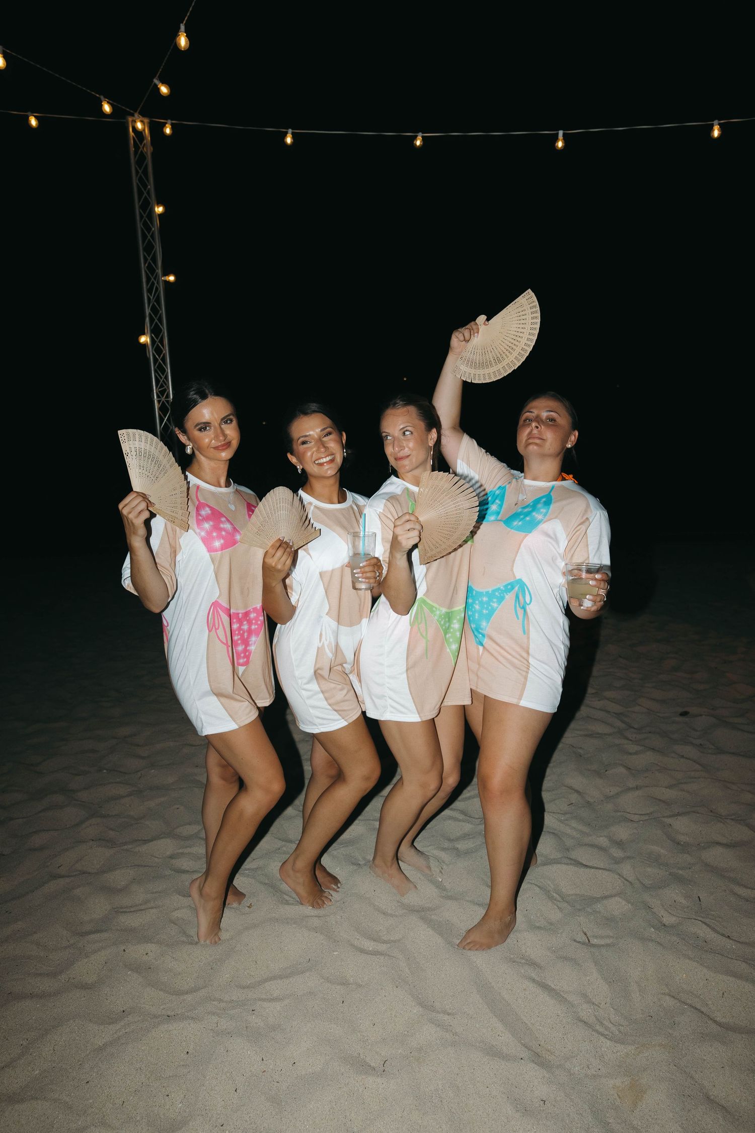 Four women posing on a beach at night holding fans, wearing white cover-ups over colorful swimsuits under string lights.