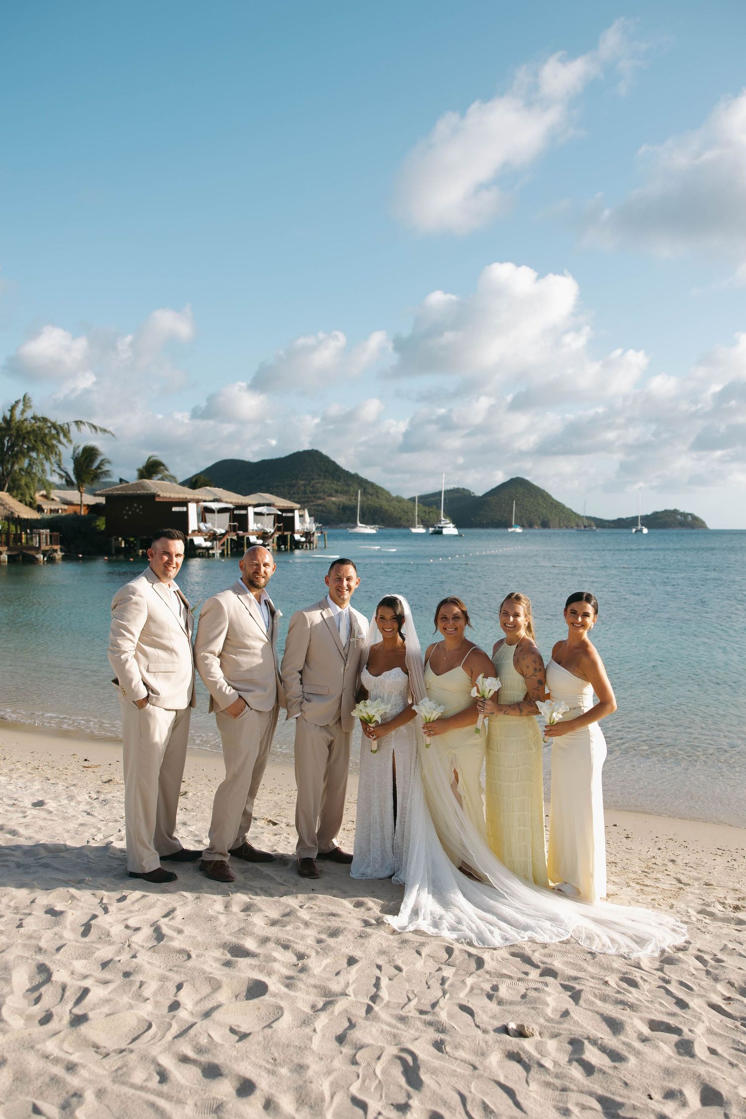 Wedding party posing on a white sand beach with turquoise water, sailboats, and tropical mountains in the background.