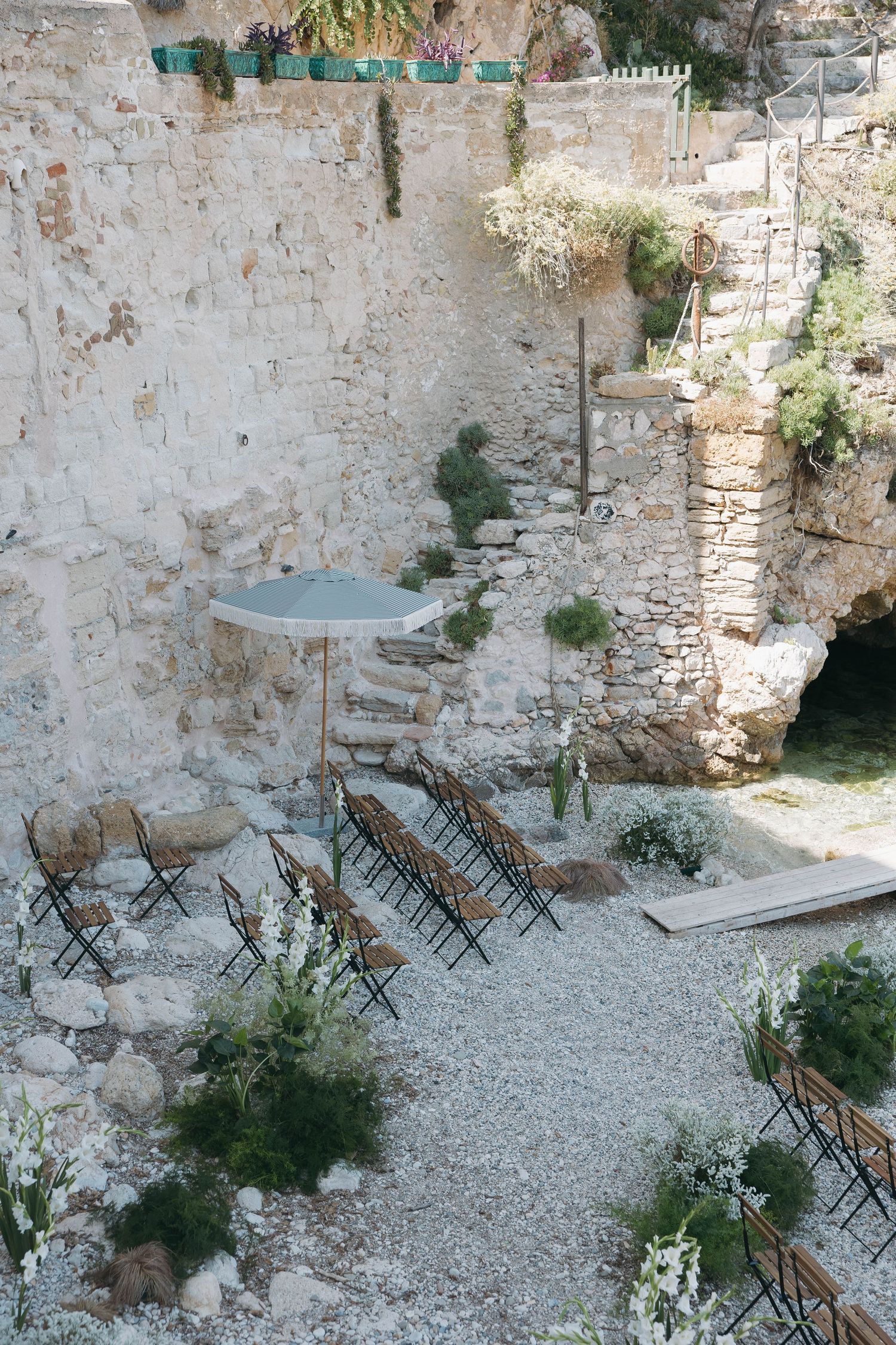 Empty wooden chairs set up in rows on pebbled ground surrounded by weathered stone walls.
