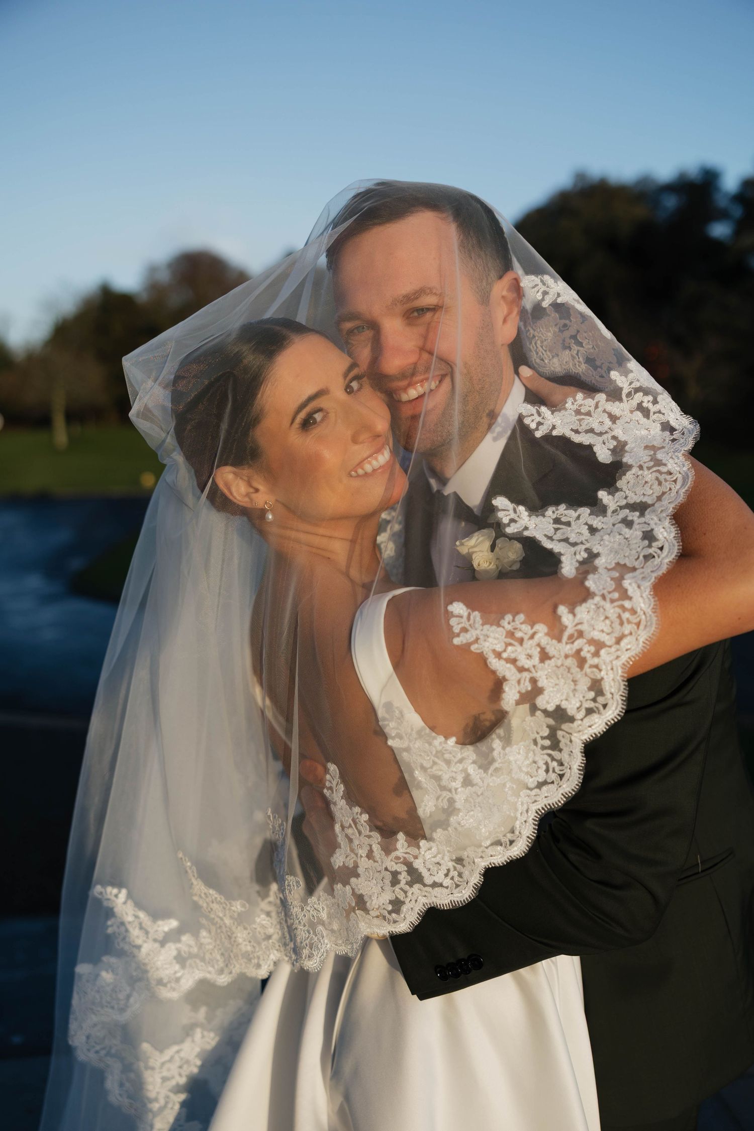 Bride and groom share intimate moment under delicate lace veil against blue sky.