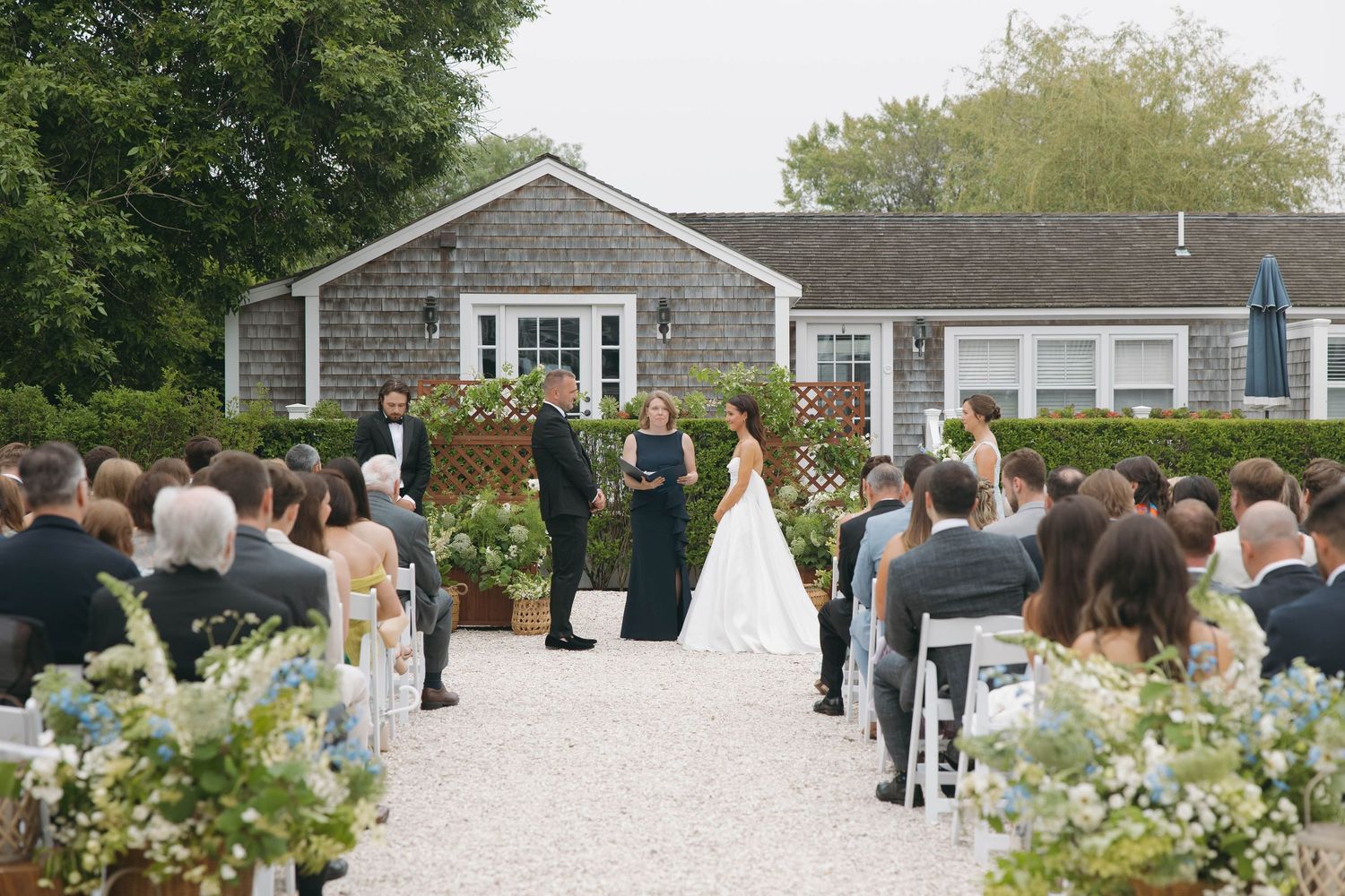 Outdoor wedding ceremony setup with white chairs and greenery against a gray shingled building.