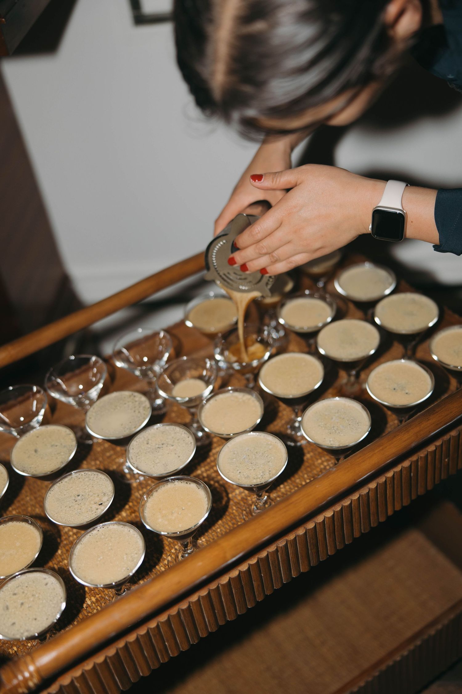 Hands pouring drinks into multiple glasses on wooden serving tray.