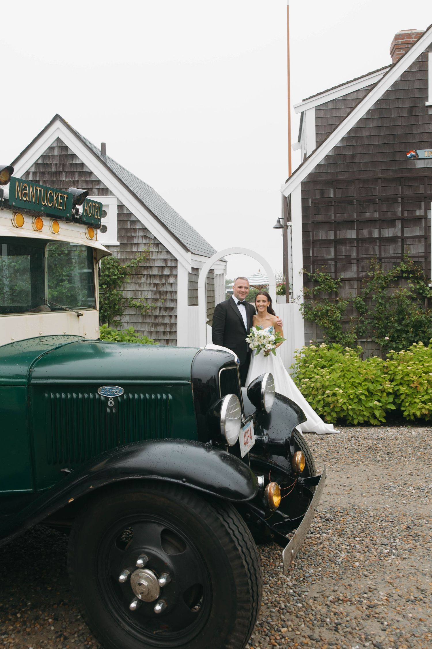 A vintage dark green car parked outside rustic shingle-style buildings on a gravel driveway.