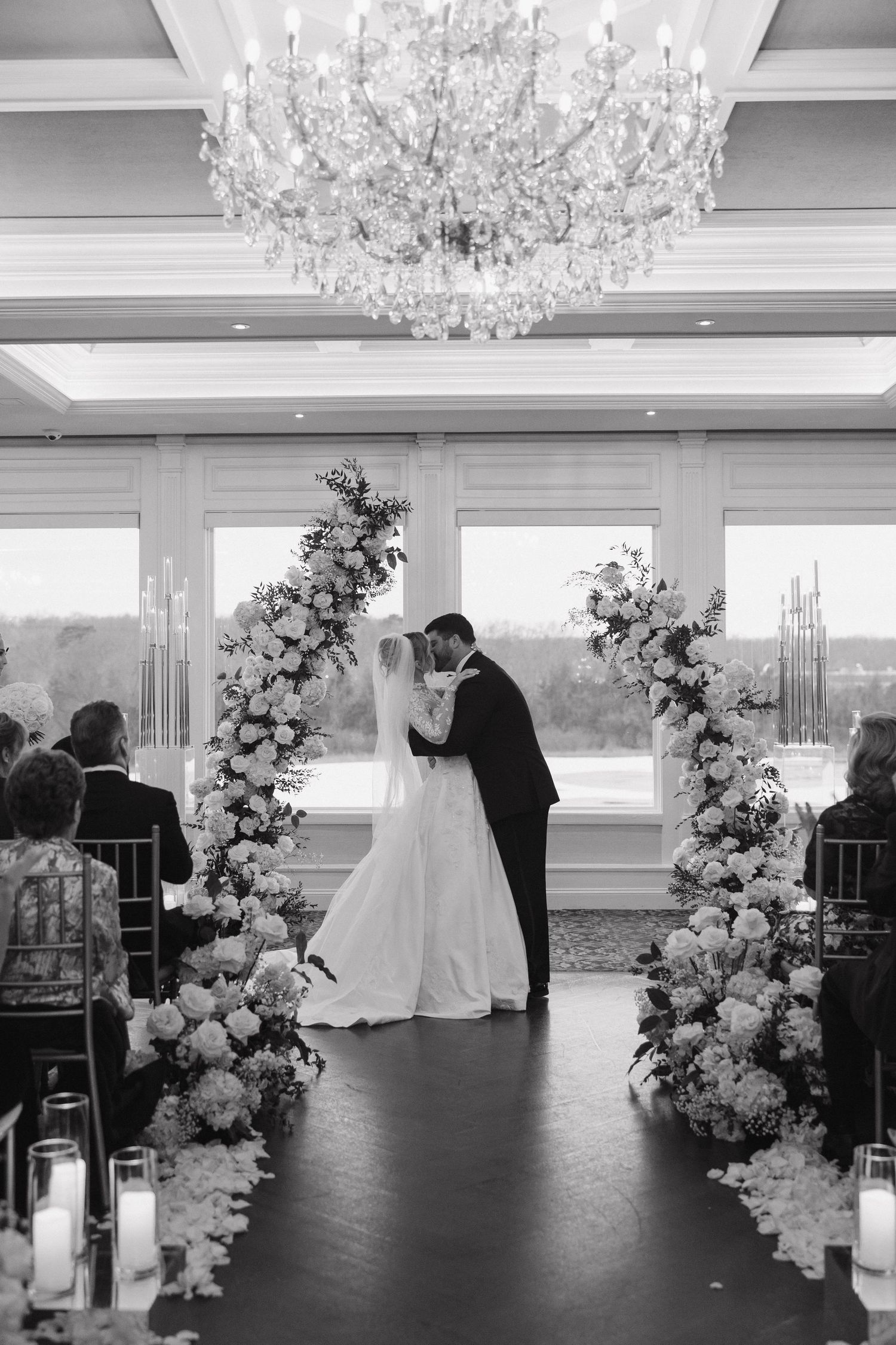 Elegant wedding ceremony with floral arch and crystal chandelier as couple shares their first kiss.