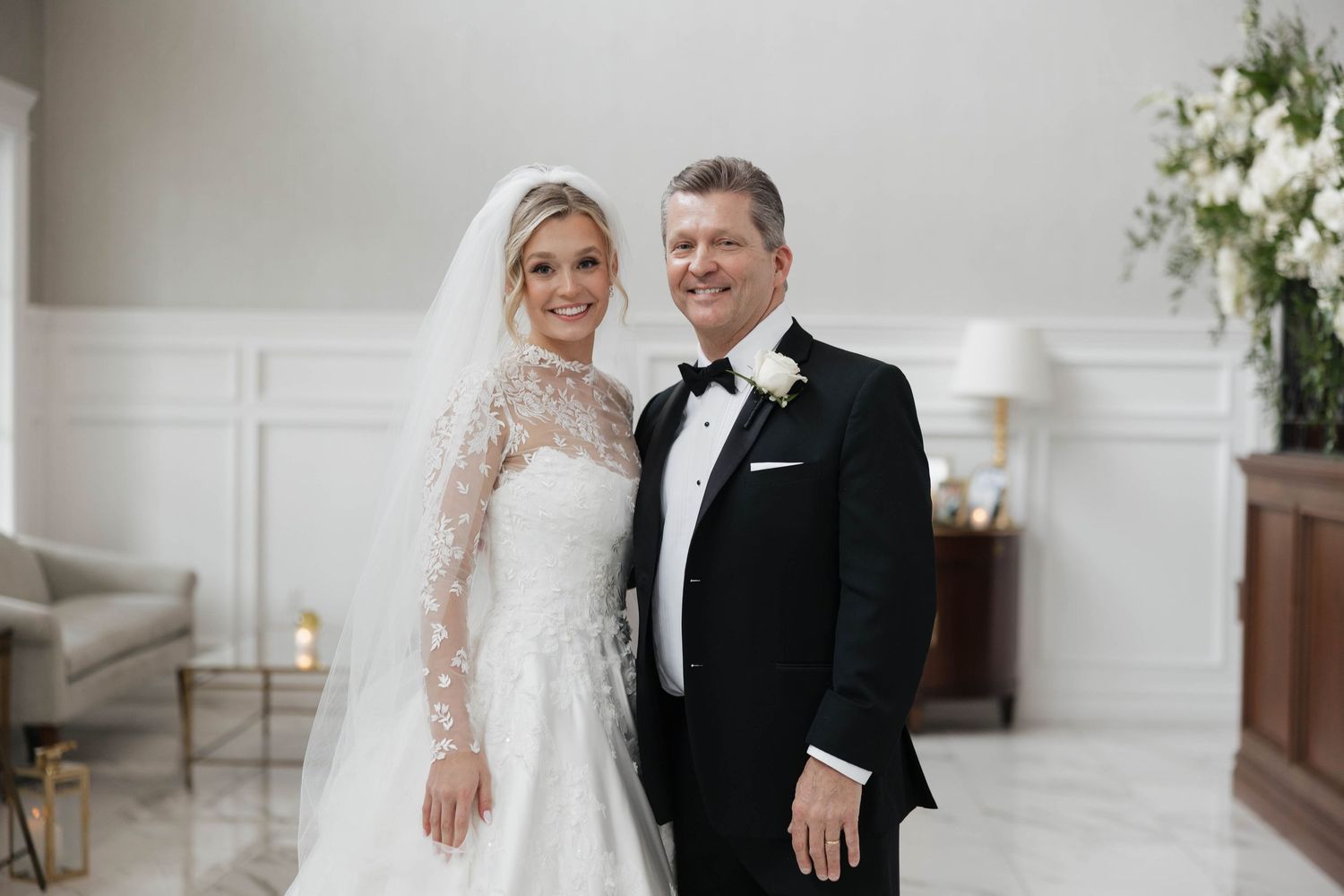 A wedding couple sharing a romantic moment in an elegant white room with floral decorations.