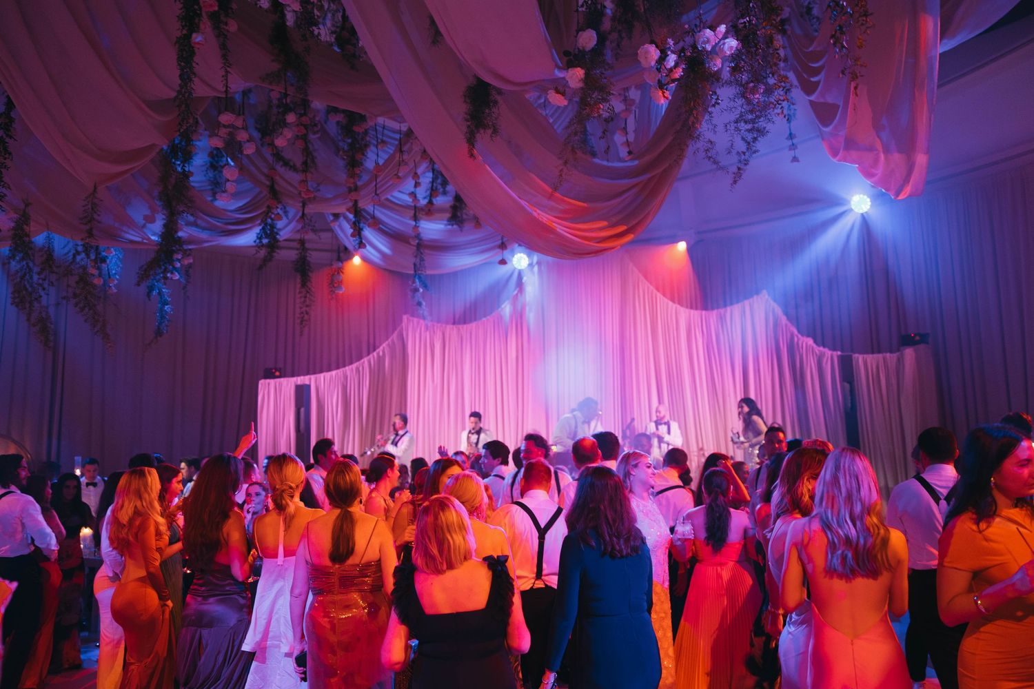 Wedding guests dance under pink and purple lighting with elegant draped ceiling decor.