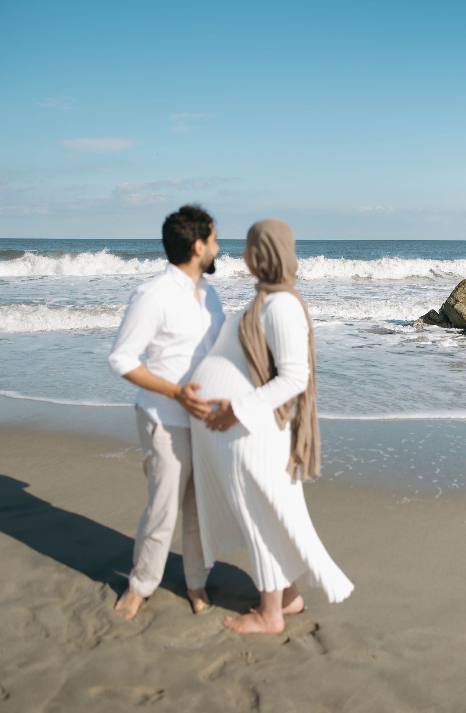 A couple in white outfits walks along a sunny beach with ocean waves crashing behind them.
