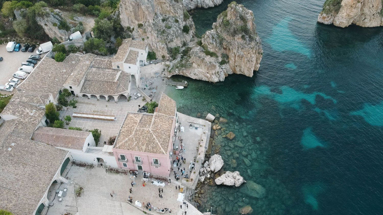 Overhead shot of coastal buildings and stone formations along crystal clear blue waters.