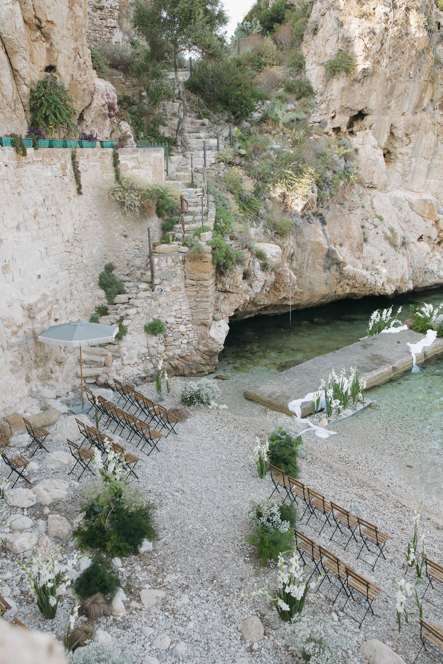 Aerial view of a coastal wedding ceremony site with folding chairs arranged on a pebble beach.