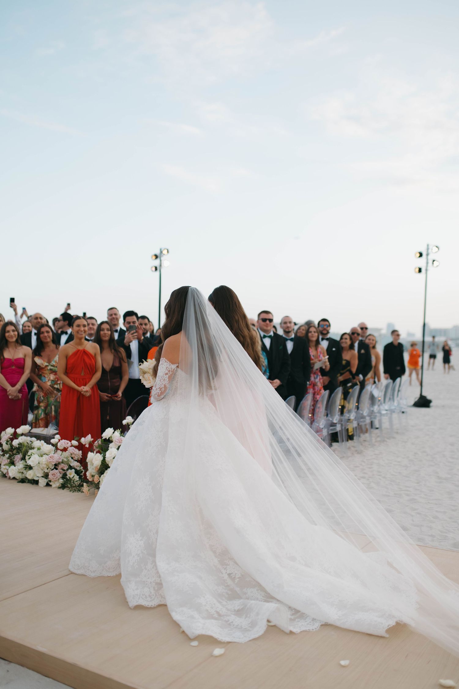 A beach wedding ceremony with flowing white gowns and guests lined up on the sand.