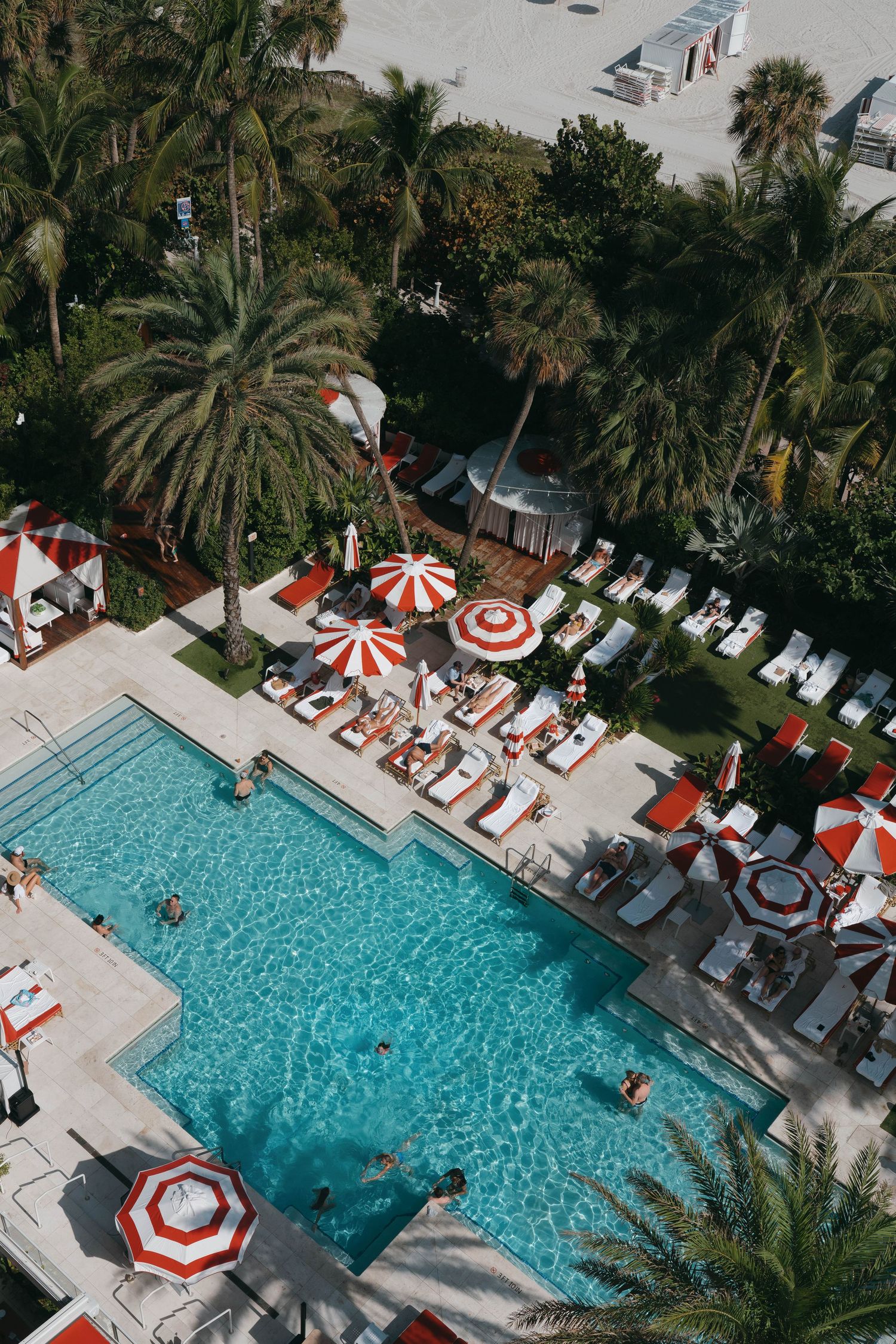 Aerial view of resort pool with red and white striped loungers surrounded by palm trees.