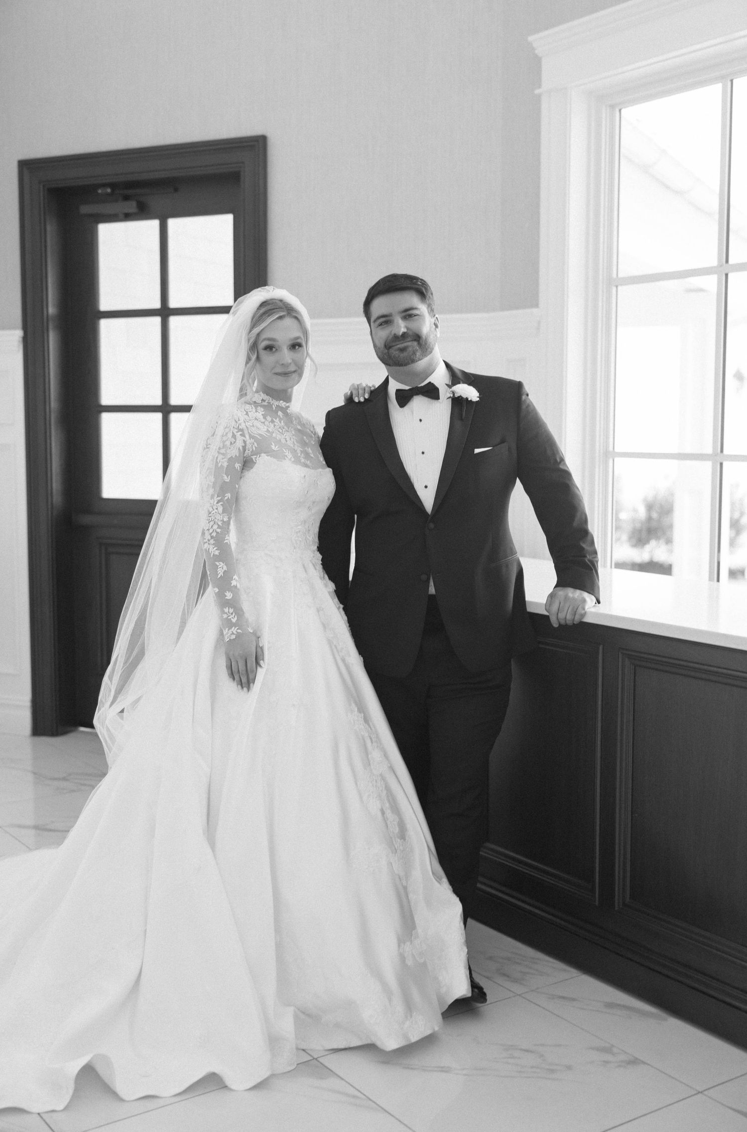 Wedding couple poses for black and white portraits in a bright interior with dark wainscoting.