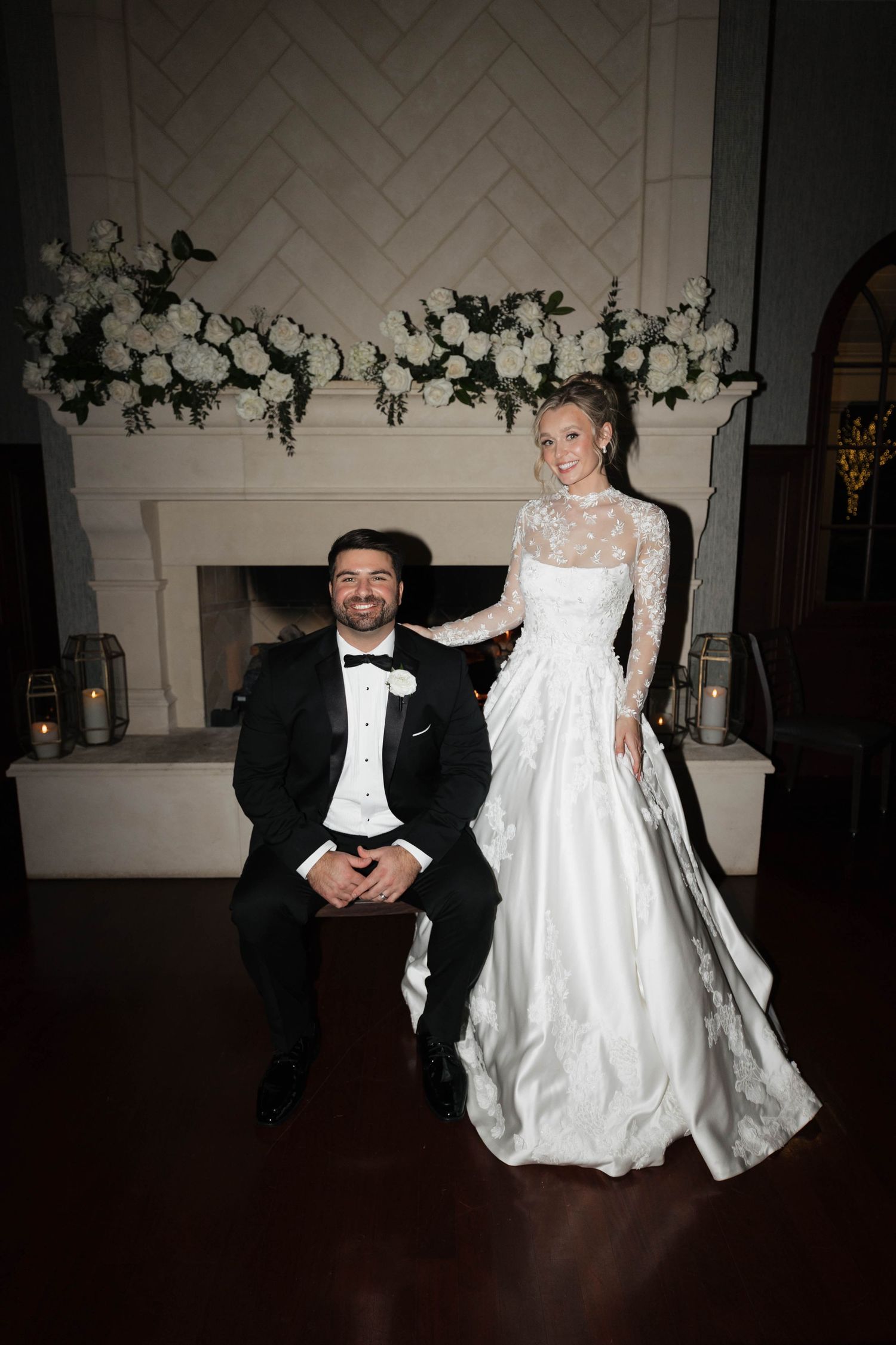 A wedding couple poses by an elegant fireplace decorated with white floral arrangements.