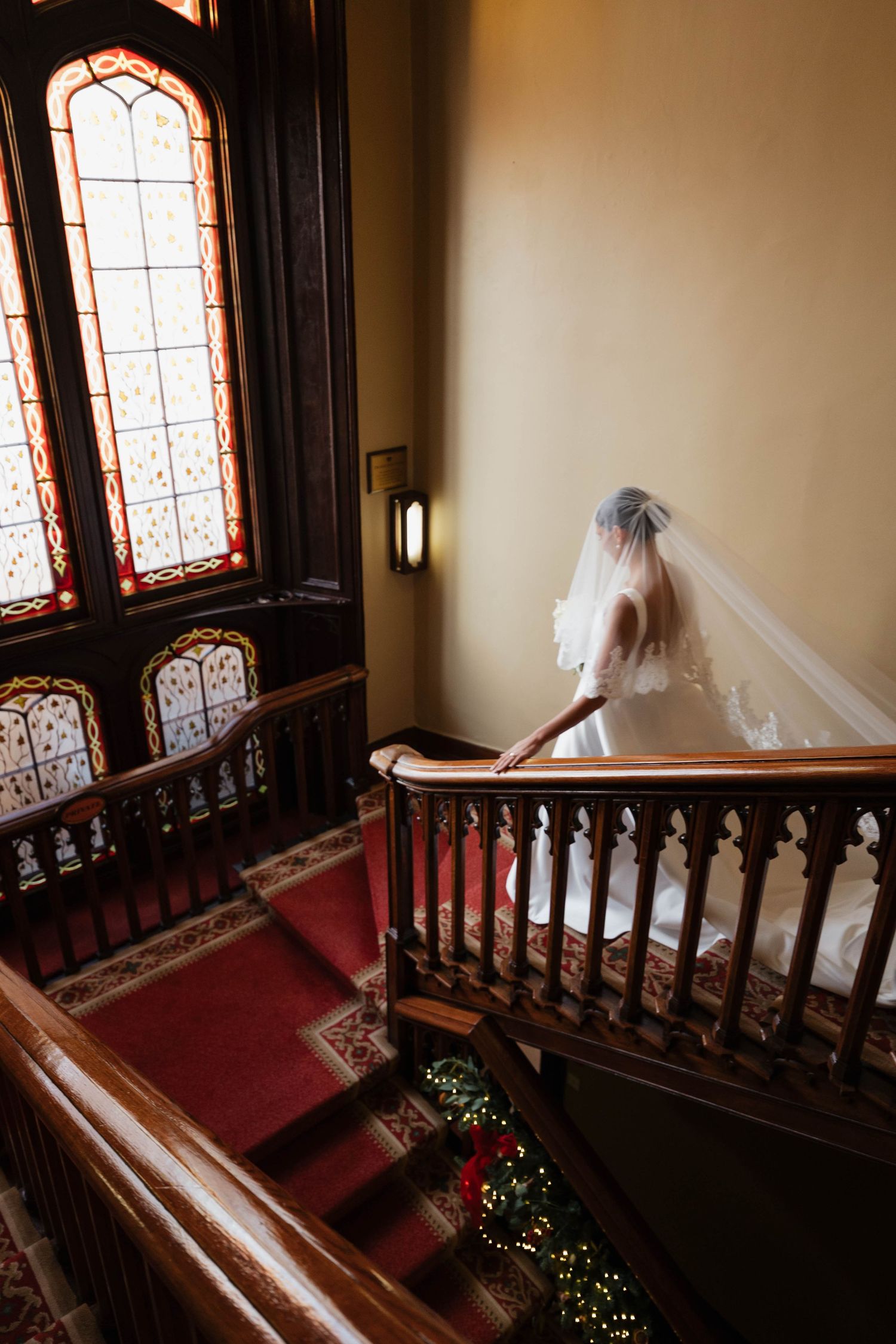 Bride in white dress and veil standing on elegant wooden staircase near stained glass windows.