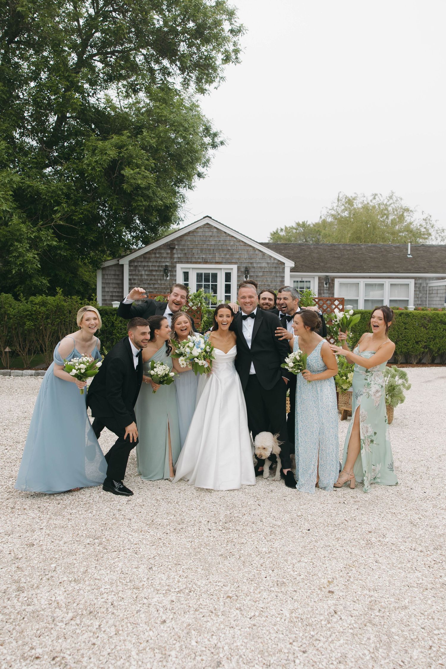 Wedding party laughs together on gravel courtyard wearing light blue dresses and black suits.
