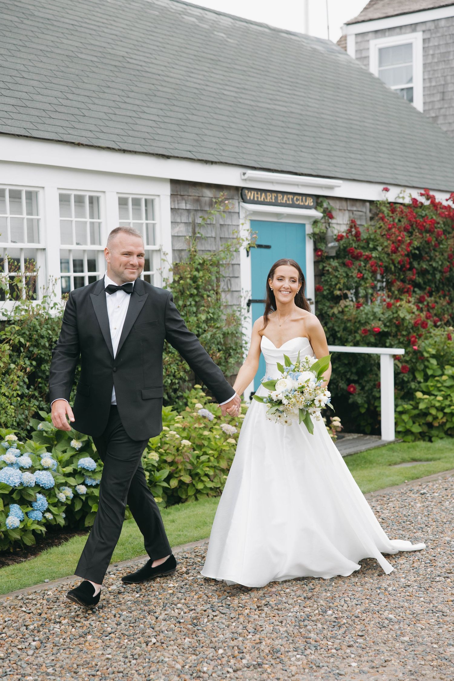 Playful wedding photos on gravel path by hydrangea garden and cottages.