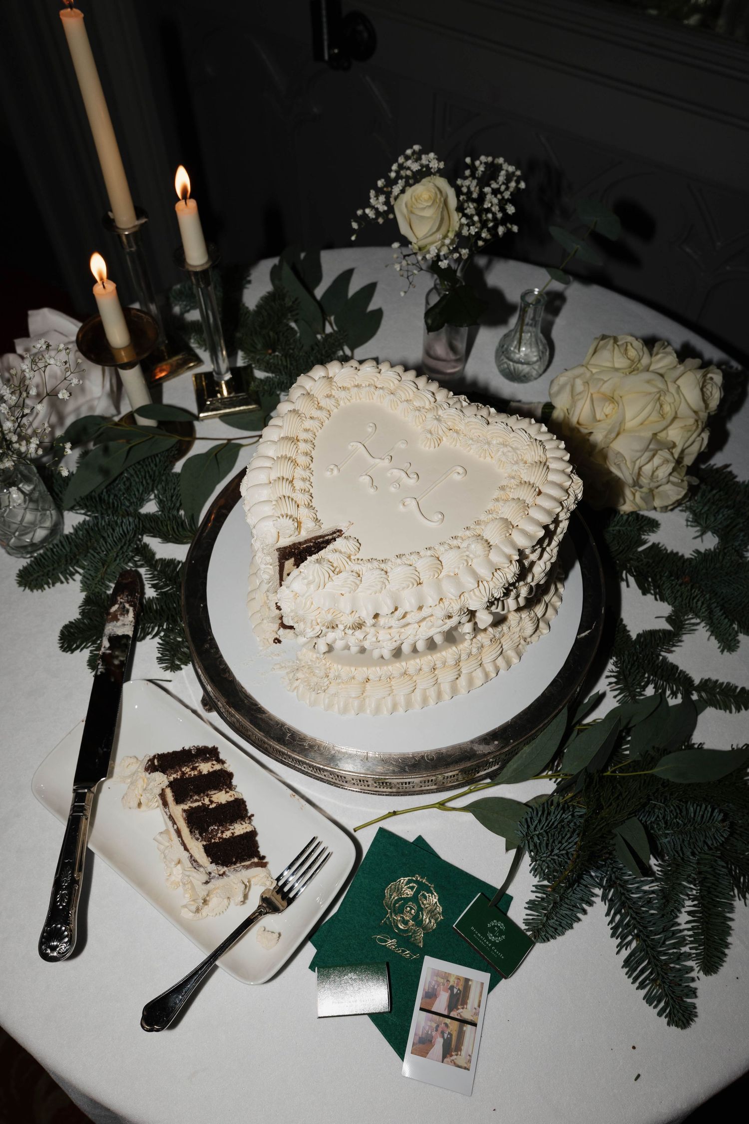 Heart-shaped wedding cake decorated in white frosting with evergreen branches and candles on elegant table setting.