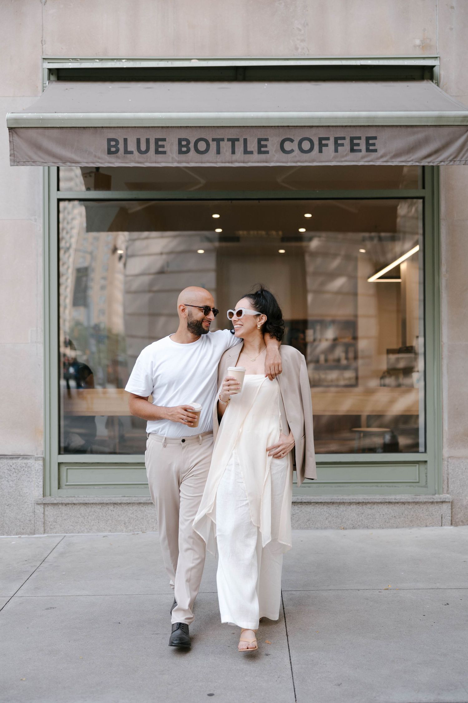 A stylish couple in white outfits walks past Blue Bottle Coffee storefront.