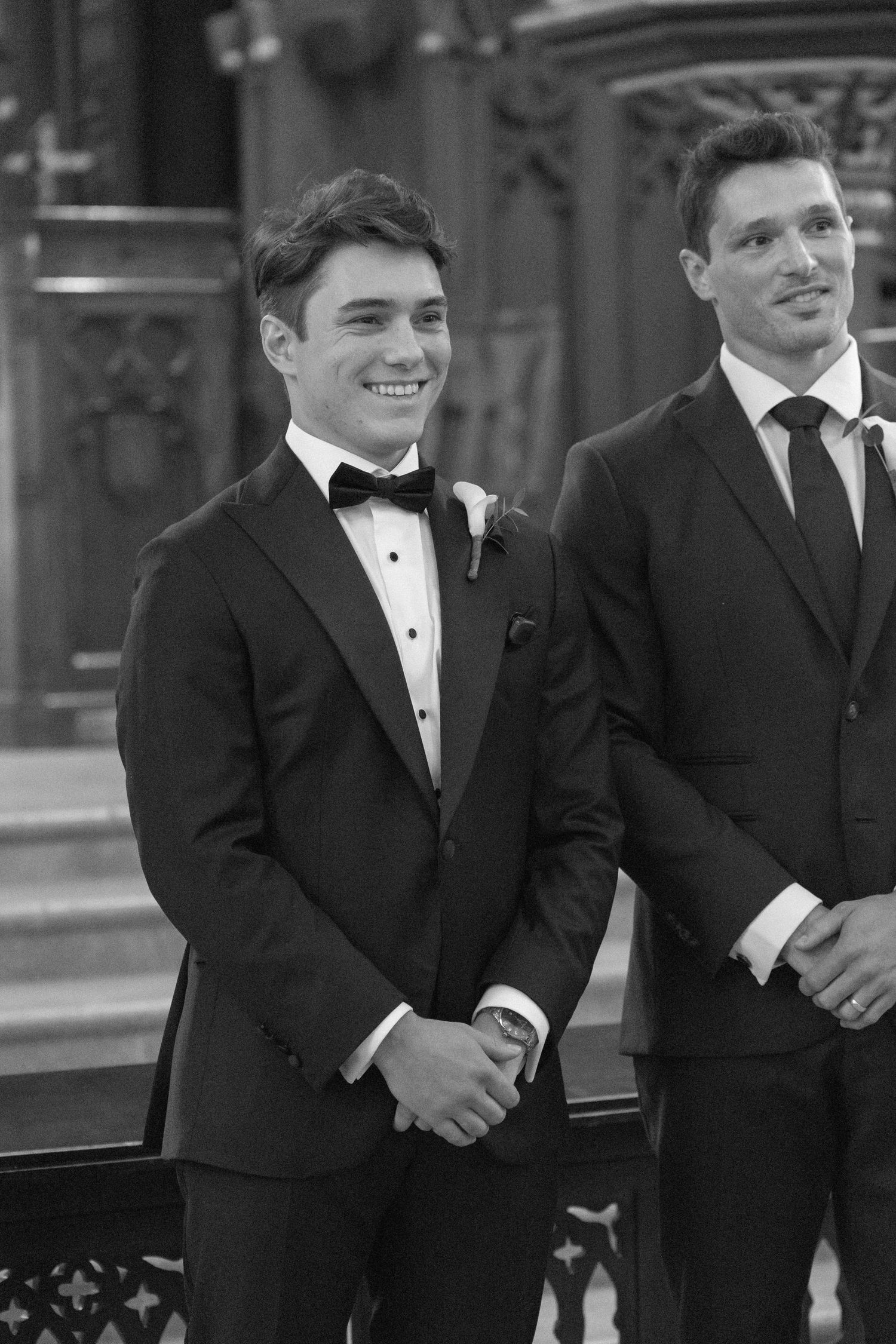 Two men in formal suits smiling at a wedding ceremony in a church, black and white photo.