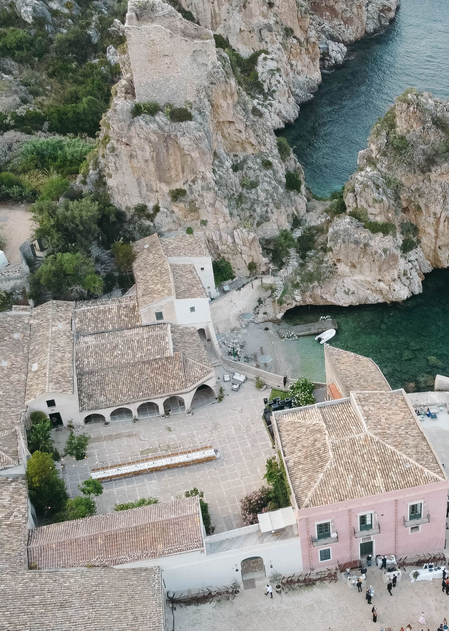 Bird's eye view of pink and beige buildings built into dramatic coastal cliffs over blue waters.