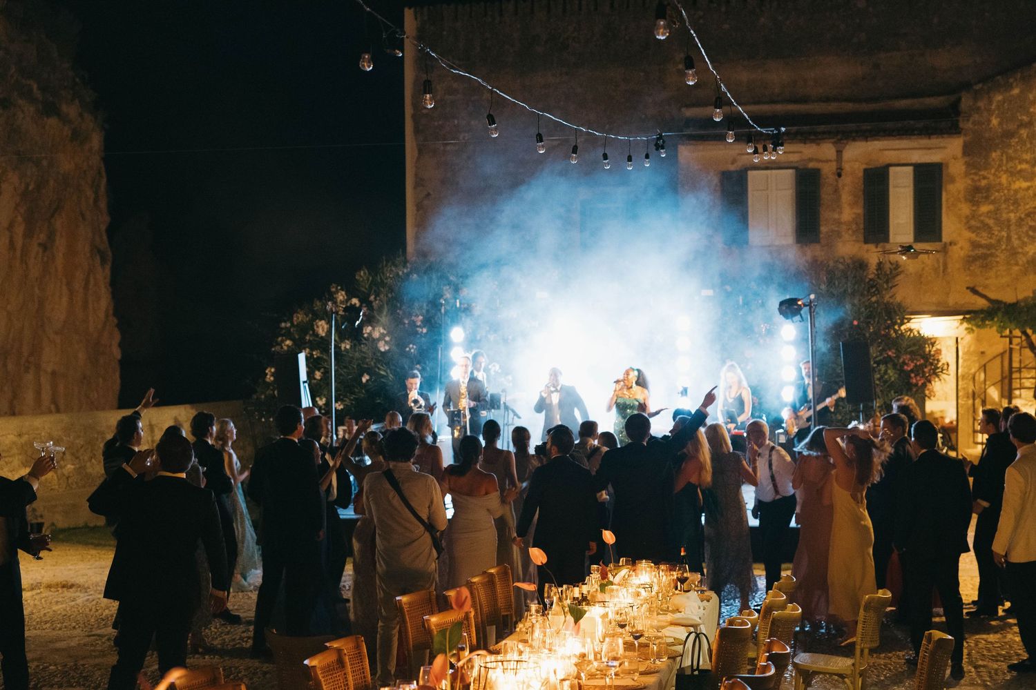 Evening party scene with blue smoke effects and string lights illuminating a stone courtyard.