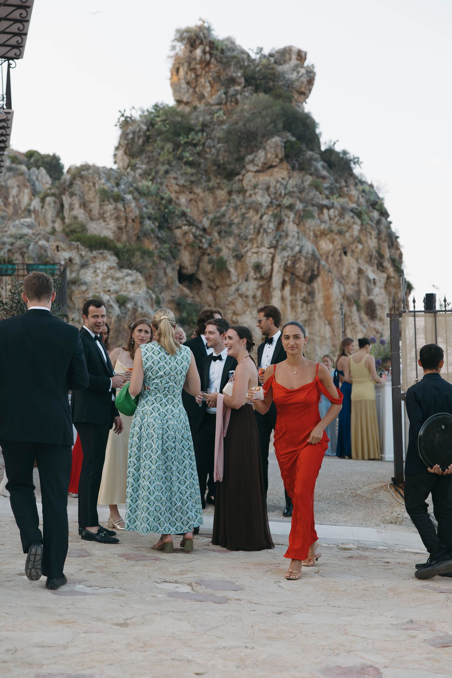 Elegant outdoor wedding reception gathering with guests mingling near rocky cliffs at sunset.