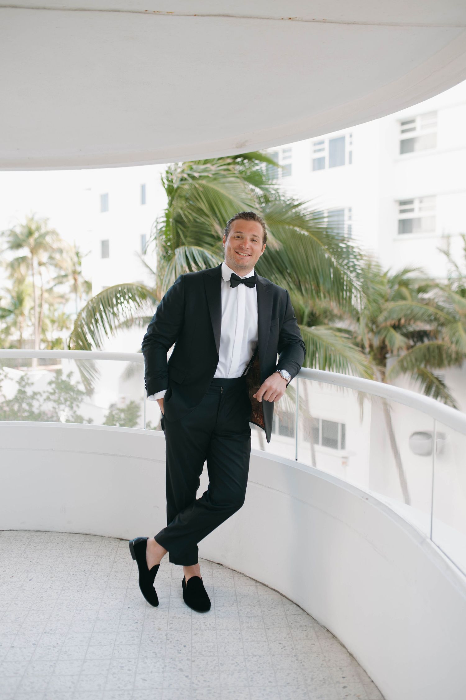 Person in a tuxedo dancing and leaping joyfully along a curved balcony with palm trees in background.