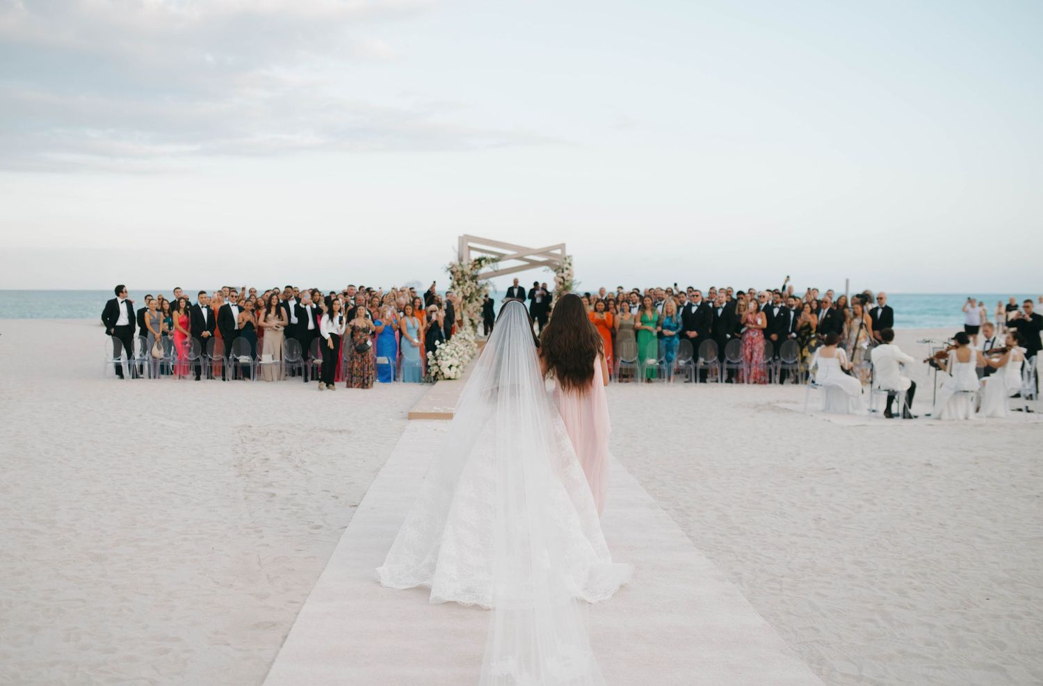 Panoramic view of a beach wedding ceremony with guests gathered on white sand.