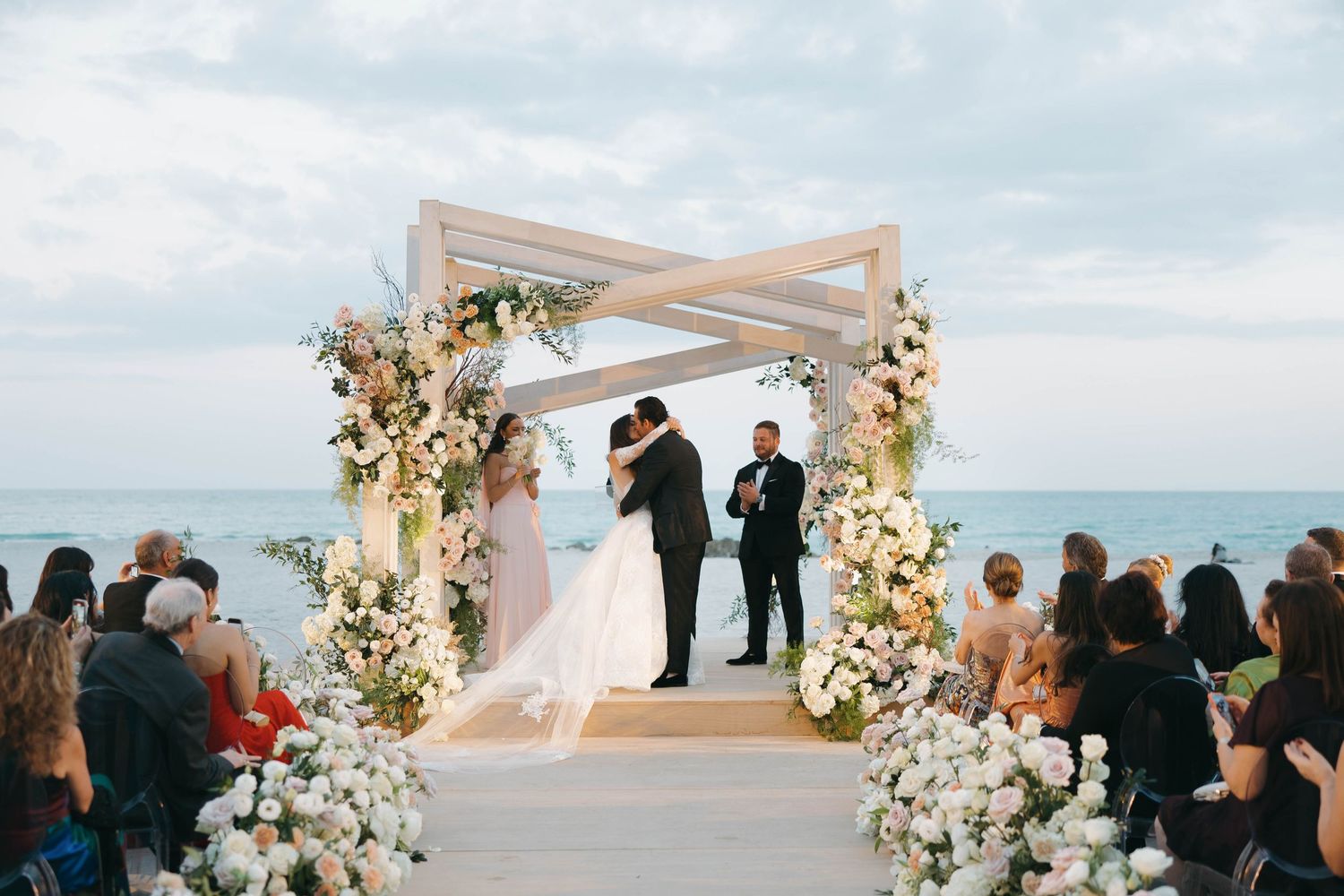 Oceanfront wedding ceremony with guests gathered around a white pergola adorned with flowers.