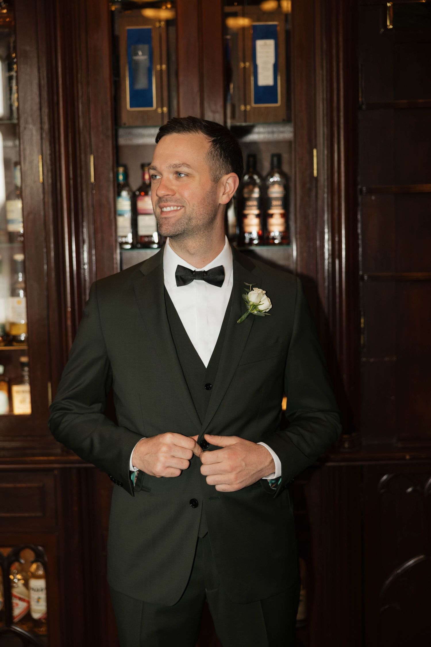 A groomsman in formal attire adjusts his suit jacket in an elegant bar setting.