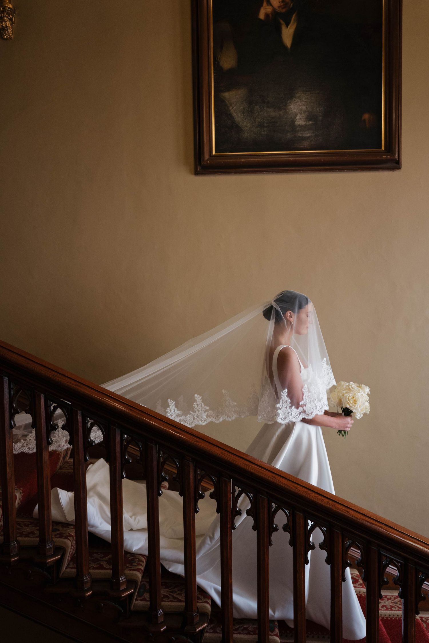 Elegant bride in flowing white dress descending a grand wooden staircase in a historic mansion.