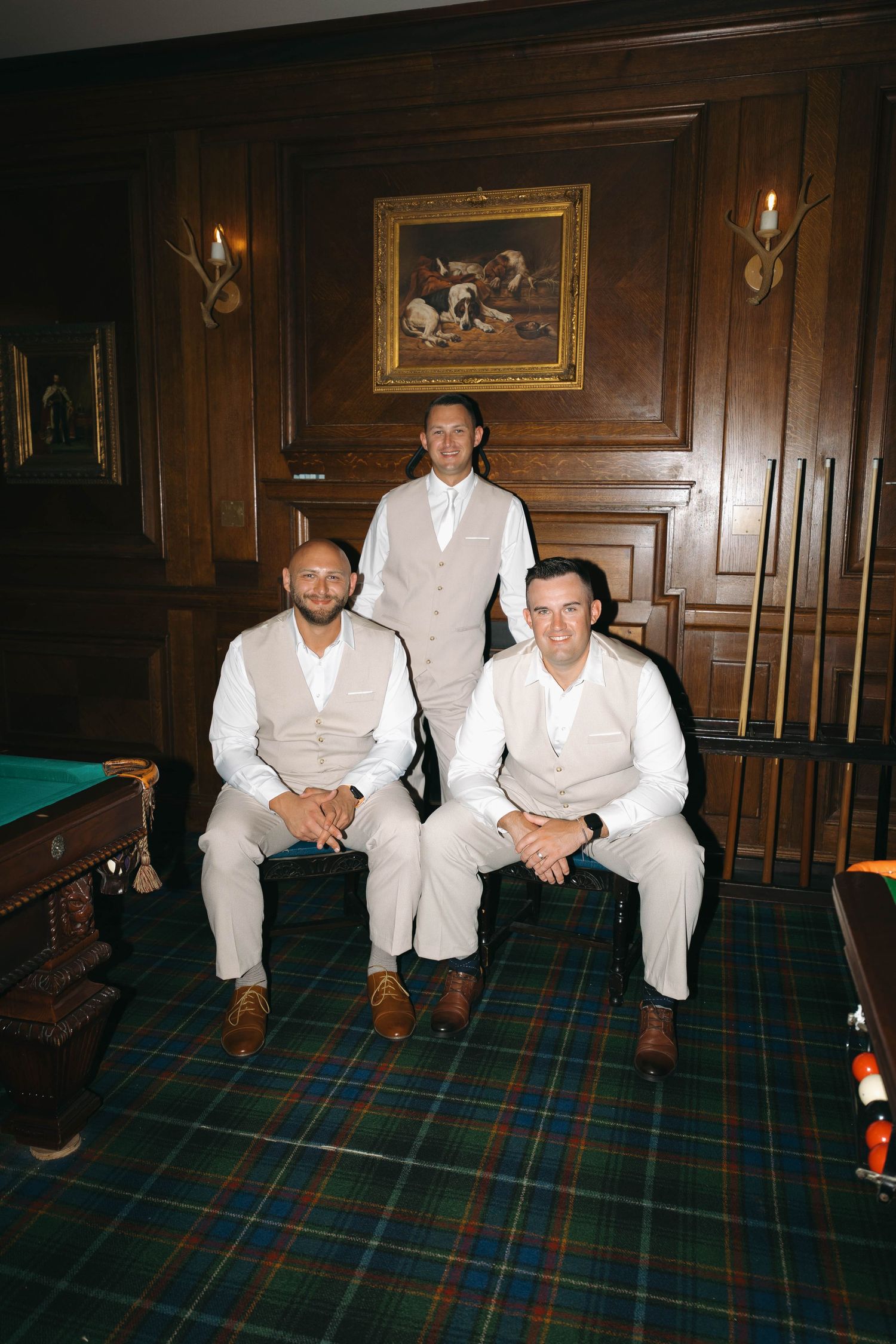 Three groomsmen in tan vests and white shirts pose in an elegant wood-paneled billiards room.