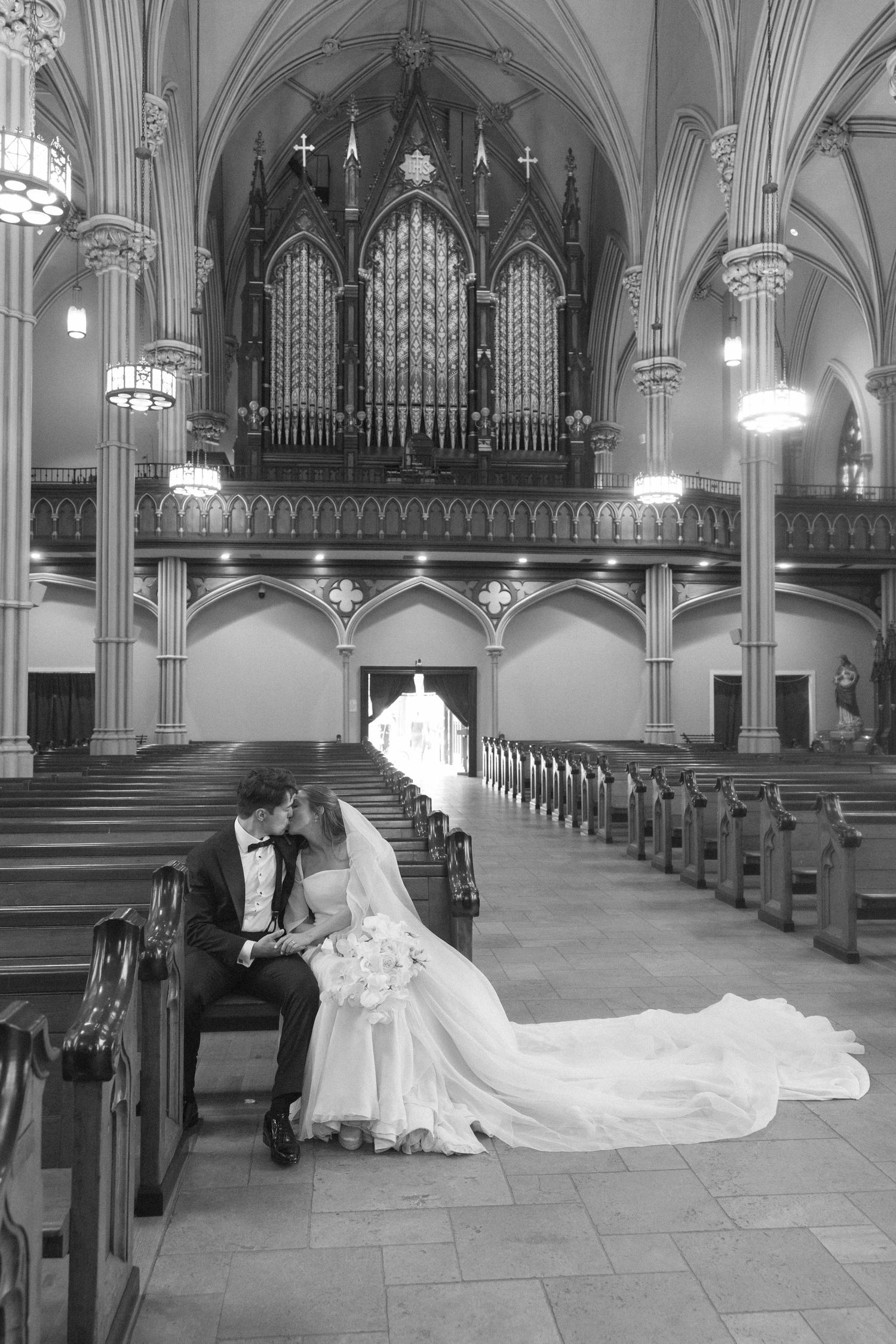 Black and white photo of bride and groom sharing a kiss on church pew with grand pipe organ backdrop.