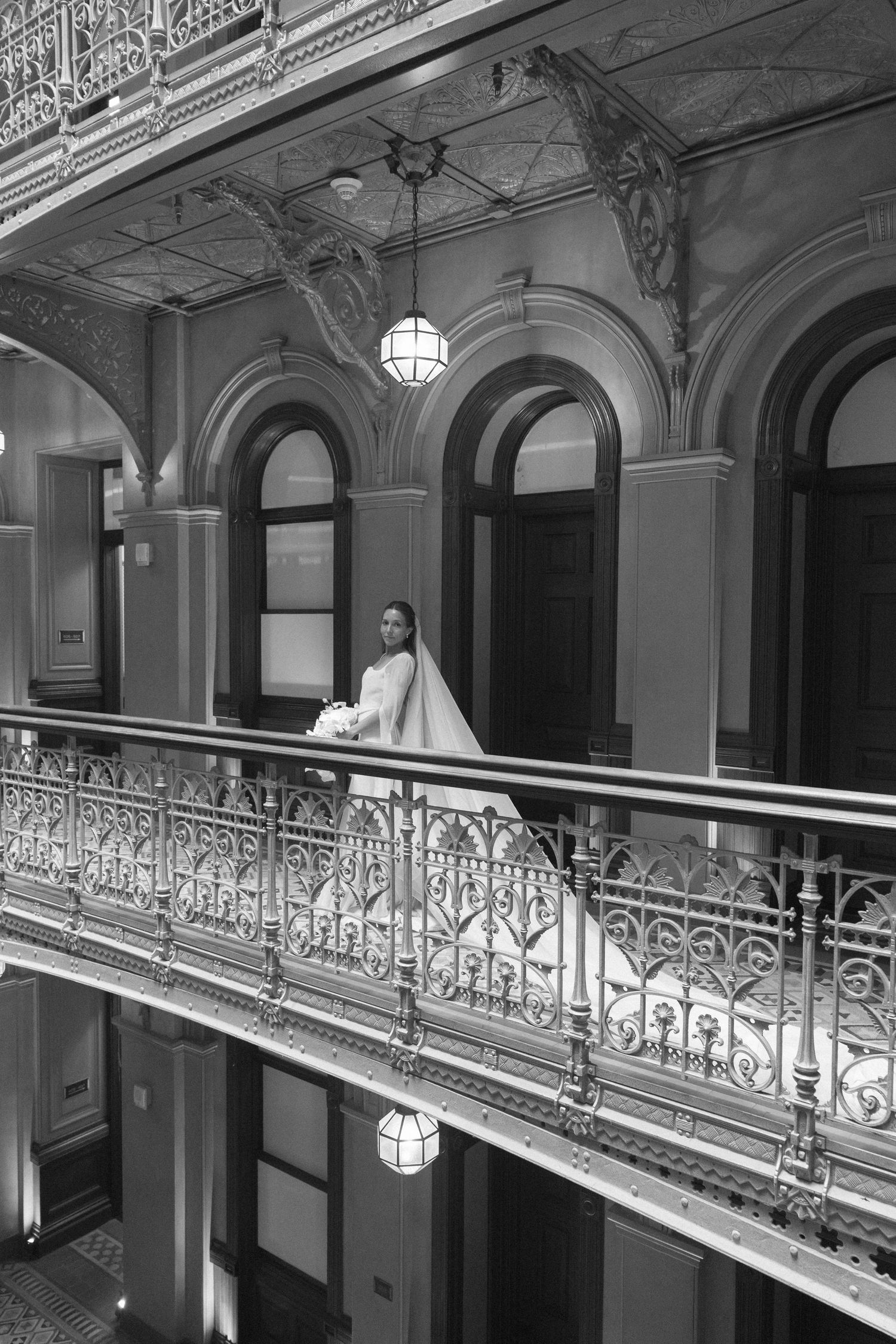 Bride in white gown stands on ornate iron balcony of elegant historic building in black and white photo.