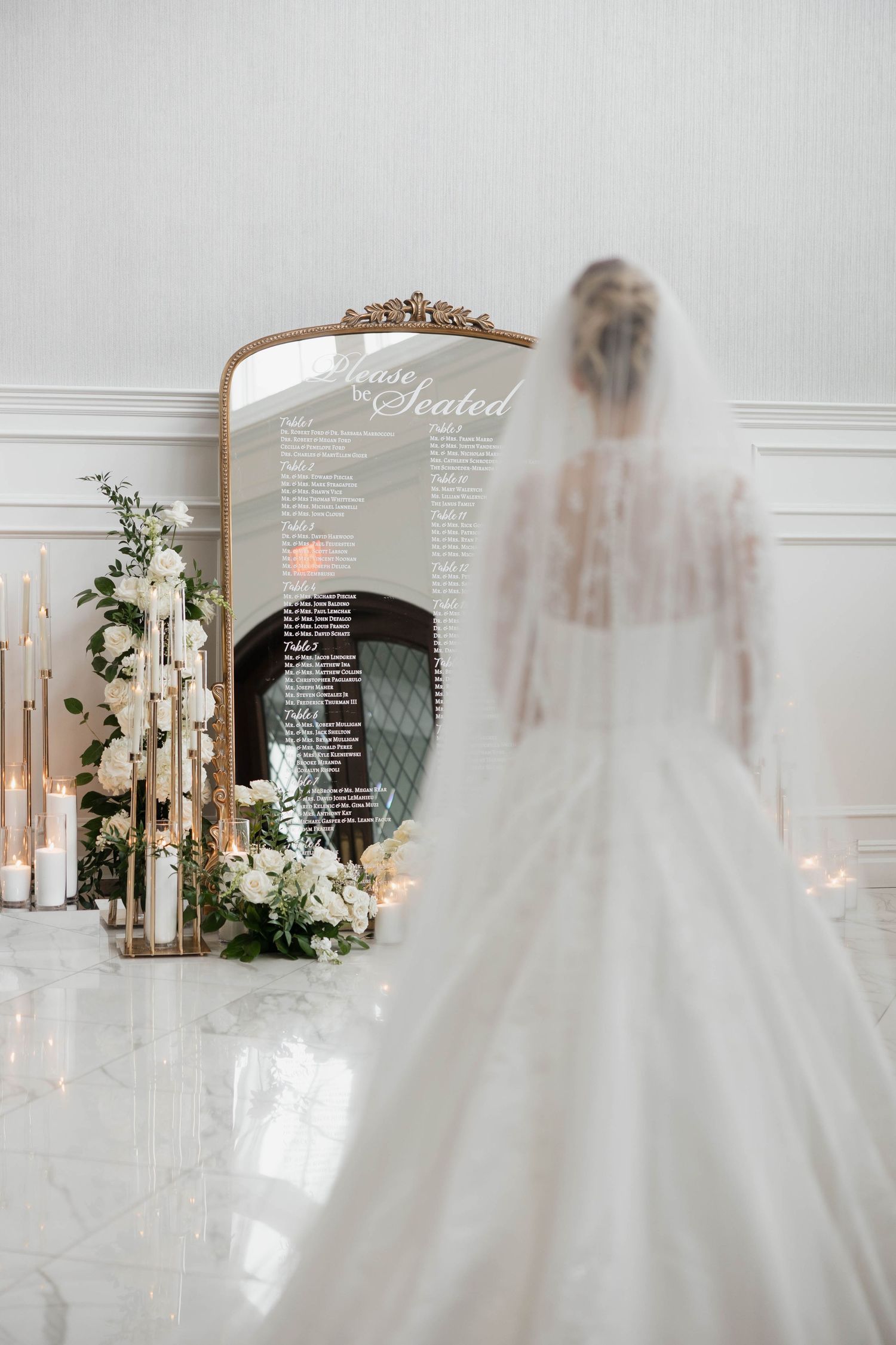 Elegant white wedding gown with flowing train displayed in a bright room with ornate mirrors and floral decor.