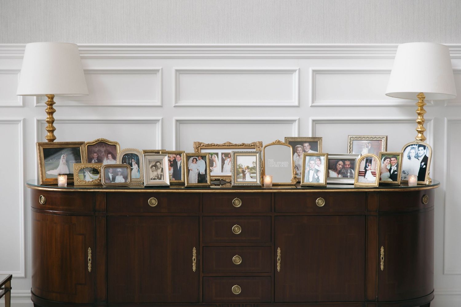 A collection of framed photographs displayed on a dark wood sideboard with white table lamps.