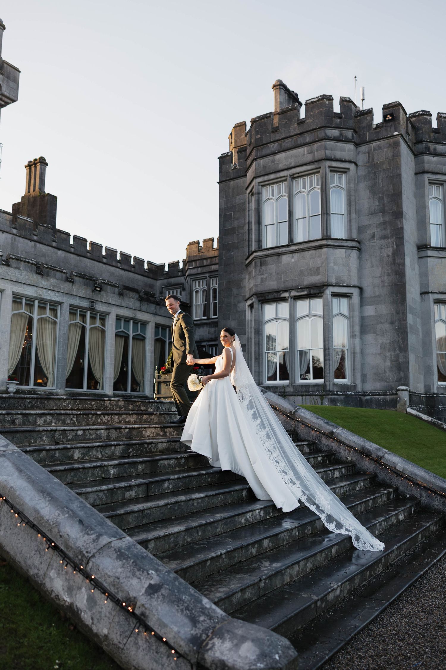 Bride in flowing white gown poses on stone steps of a historic castle.