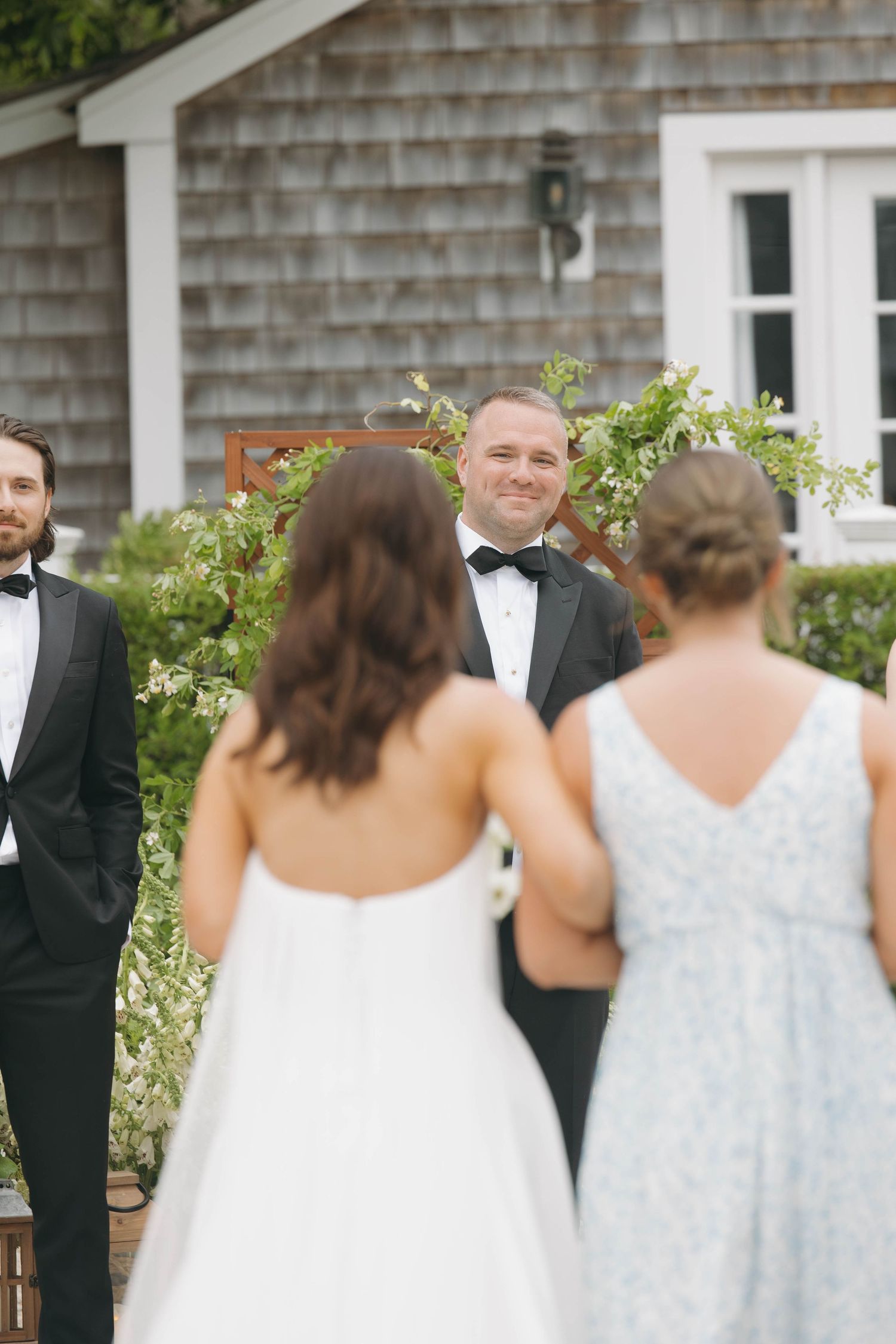 Wedding party members in formal attire gather outdoors at a rustic shingle-sided venue.