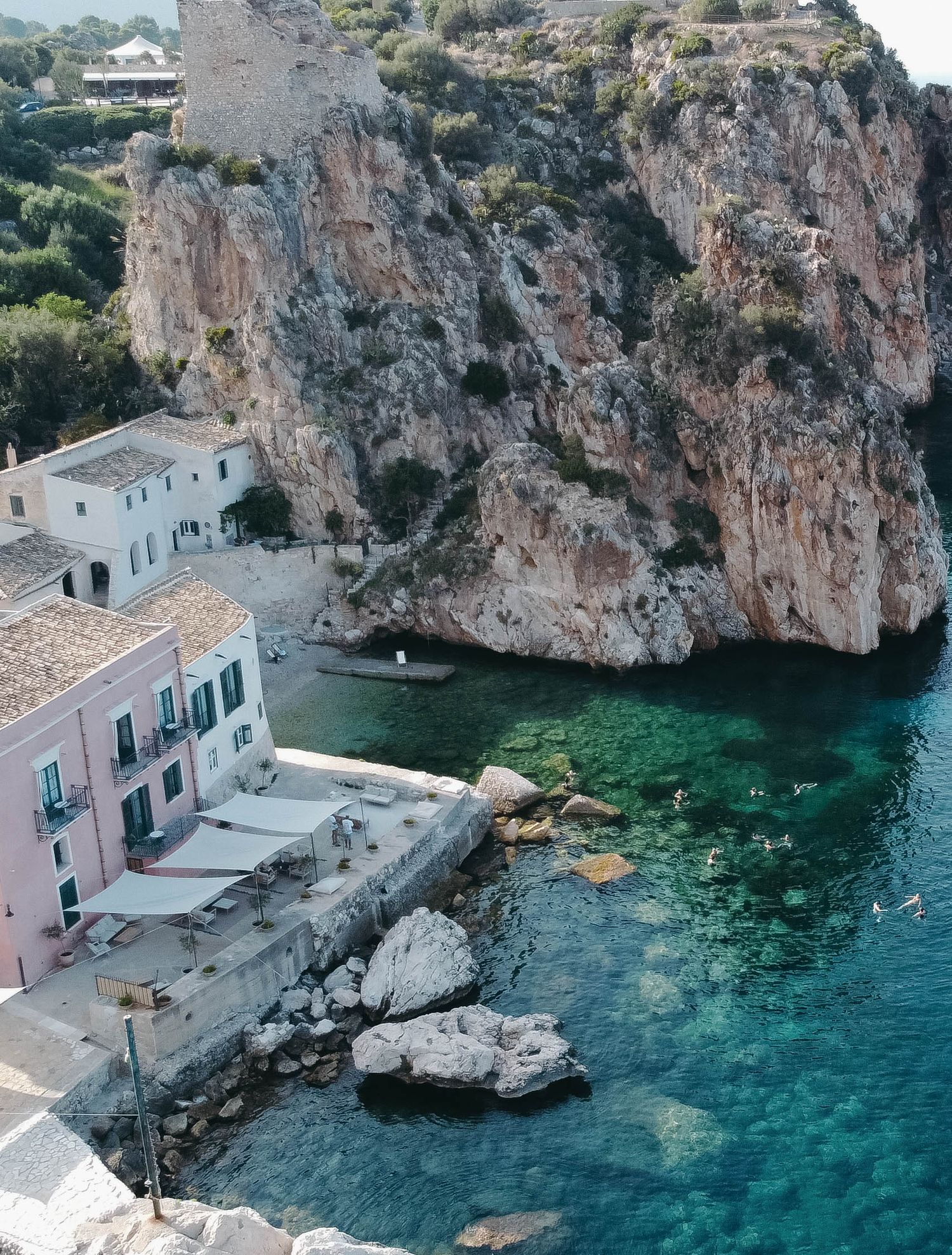 Aerial view of white buildings nestled into coastal cliffs above crystal clear Mediterranean waters.