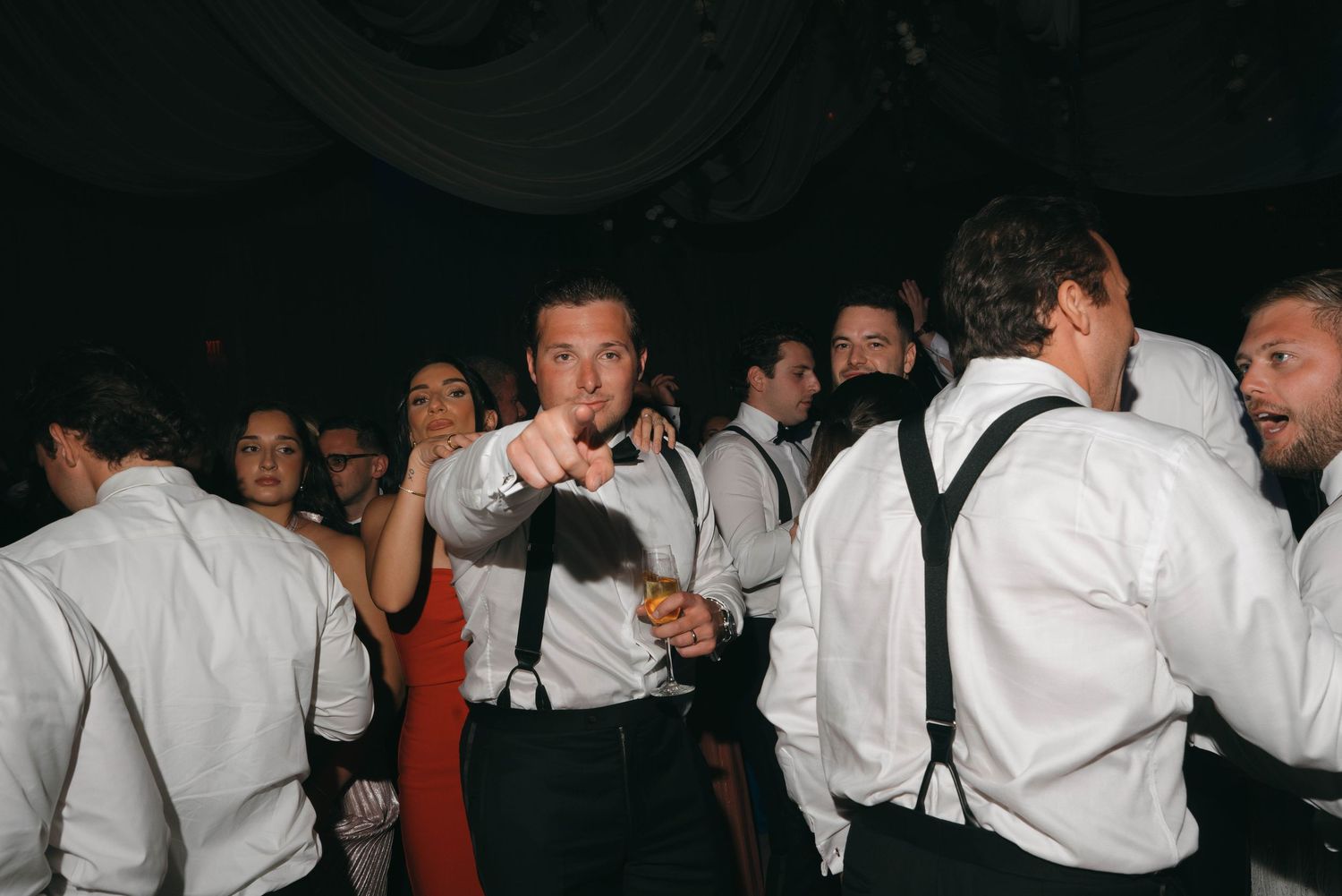 Wedding guests in formal attire dancing and celebrating in a dark reception space.